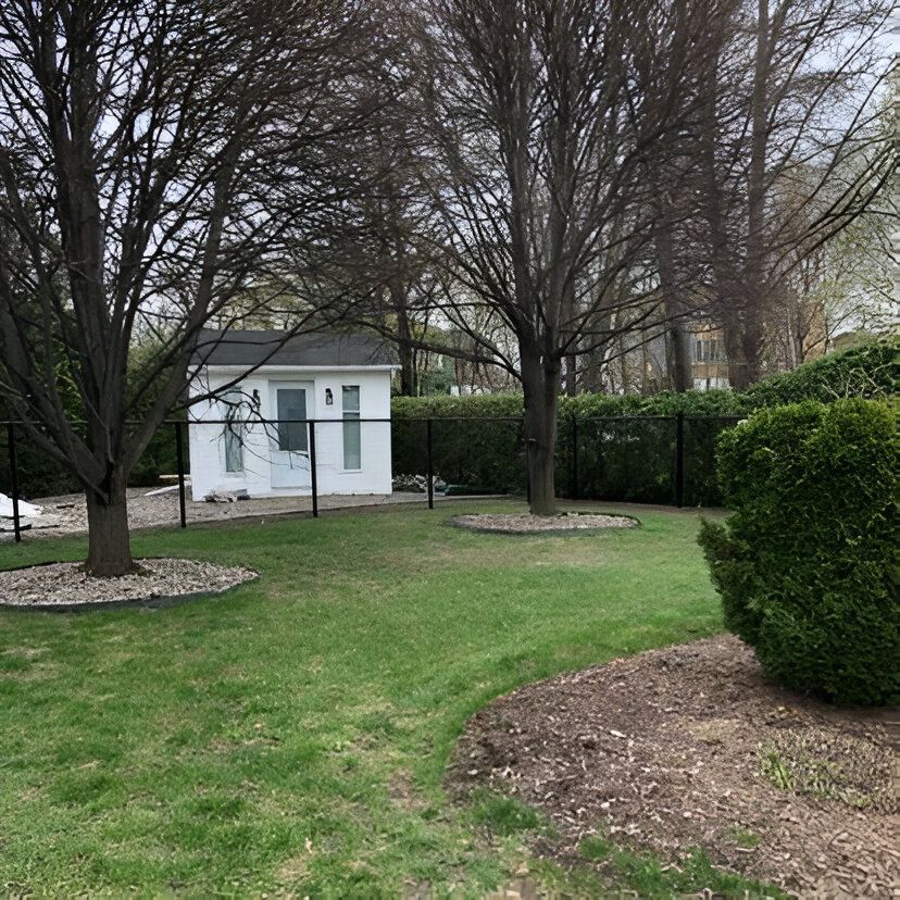 Cabanon blanc dans un jardin clôturé, arboré et verdoyant. Ciel nuageux.