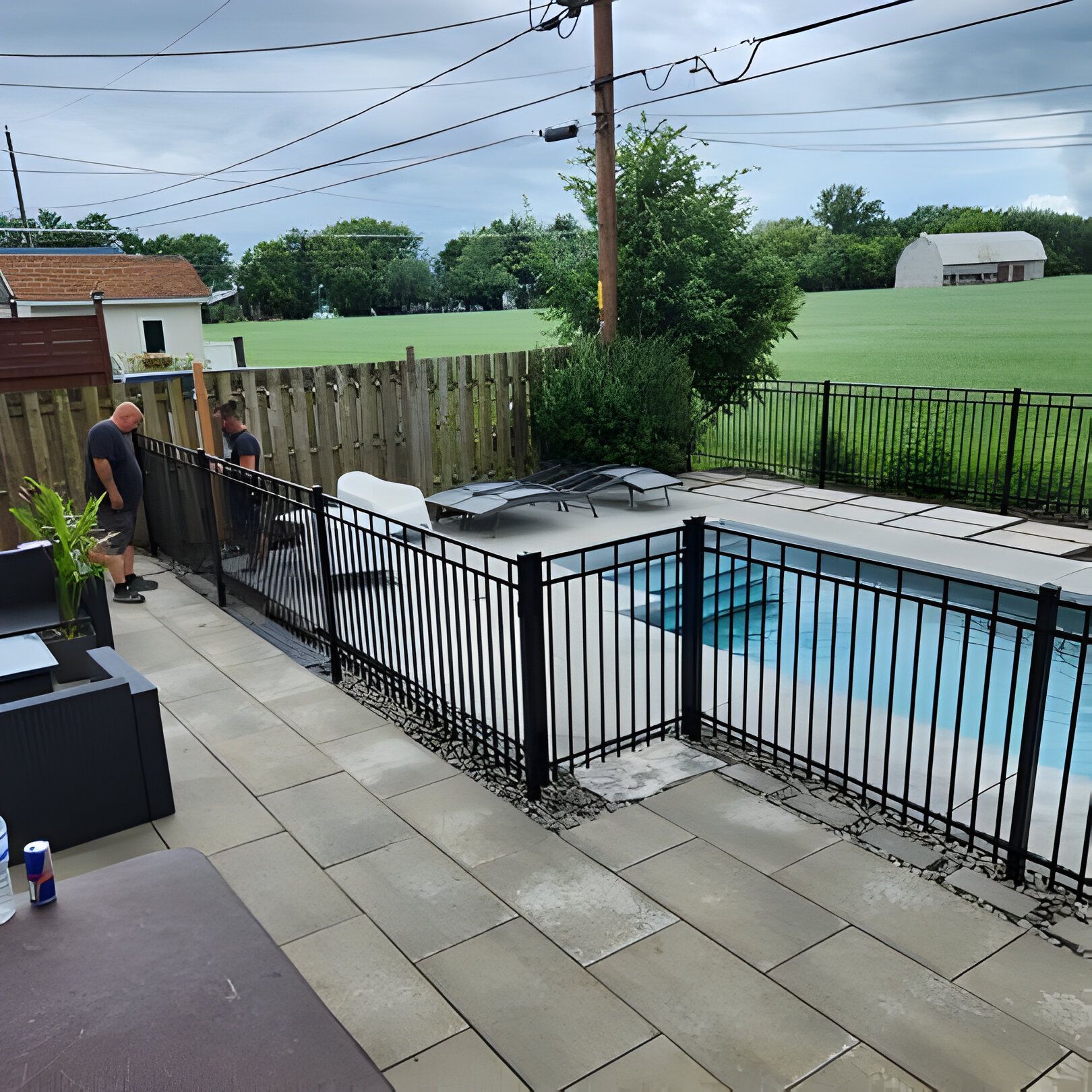 Piscine clôturée en noir avec terrasse, deux hommes et arrière-plan herbeux.