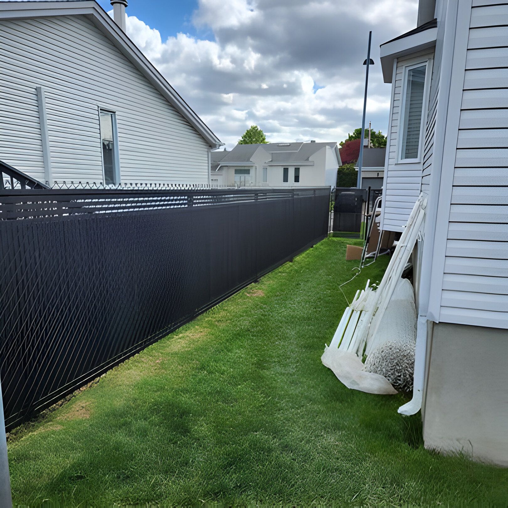 Une clôture en grillage noir sépare des jardins verdoyants entre deux maisons blanches sous un ciel nuageux.