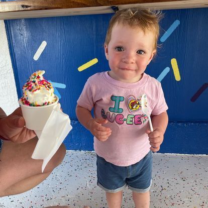 A little girl is standing next to a person holding a cup of ice cream.