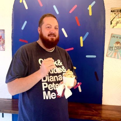 A man with a beard is holding a cup of ice cream in front of a wall with sprinkles on it