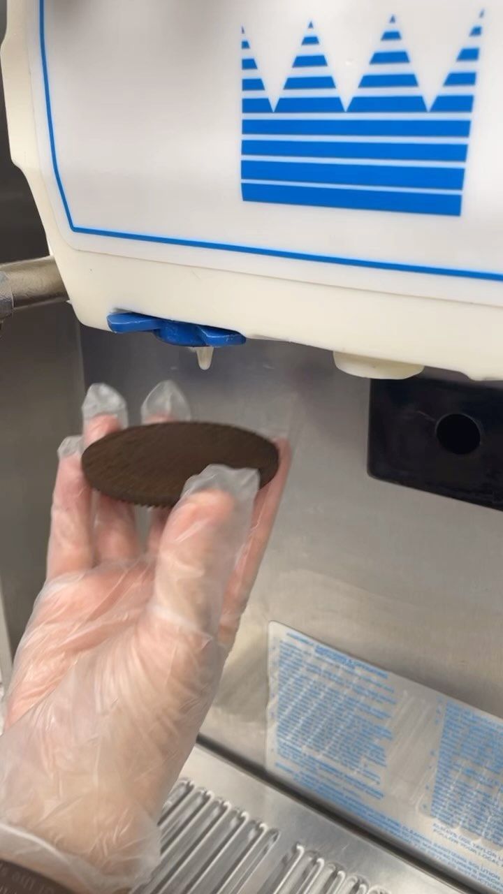 A person wearing gloves is holding an oreo cookie in front of an ice cream machine.