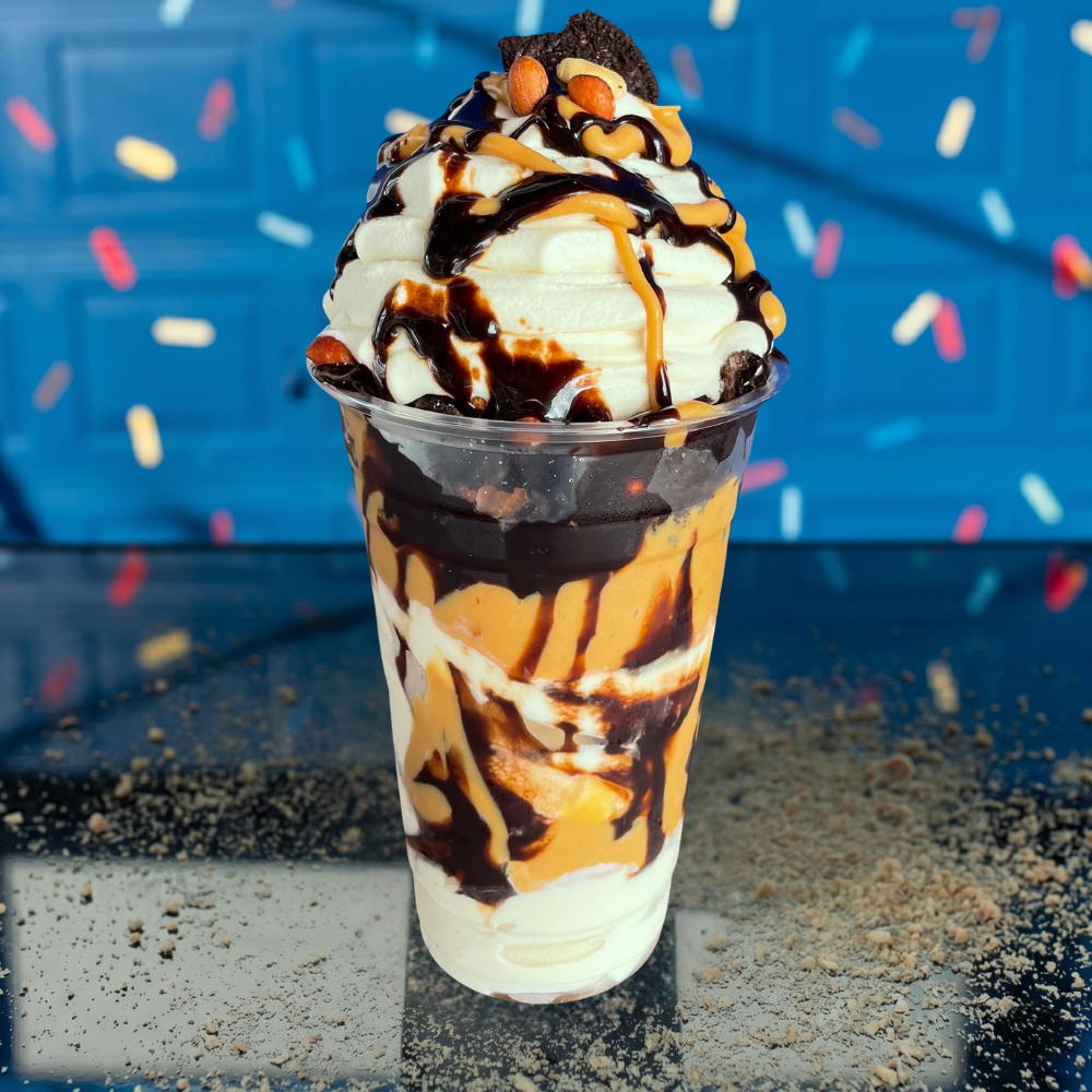 A clear plastic cup of layered vanilla ice cream, peanut butter, and chocolate sauce, topped with M&Ms, on a blue table.