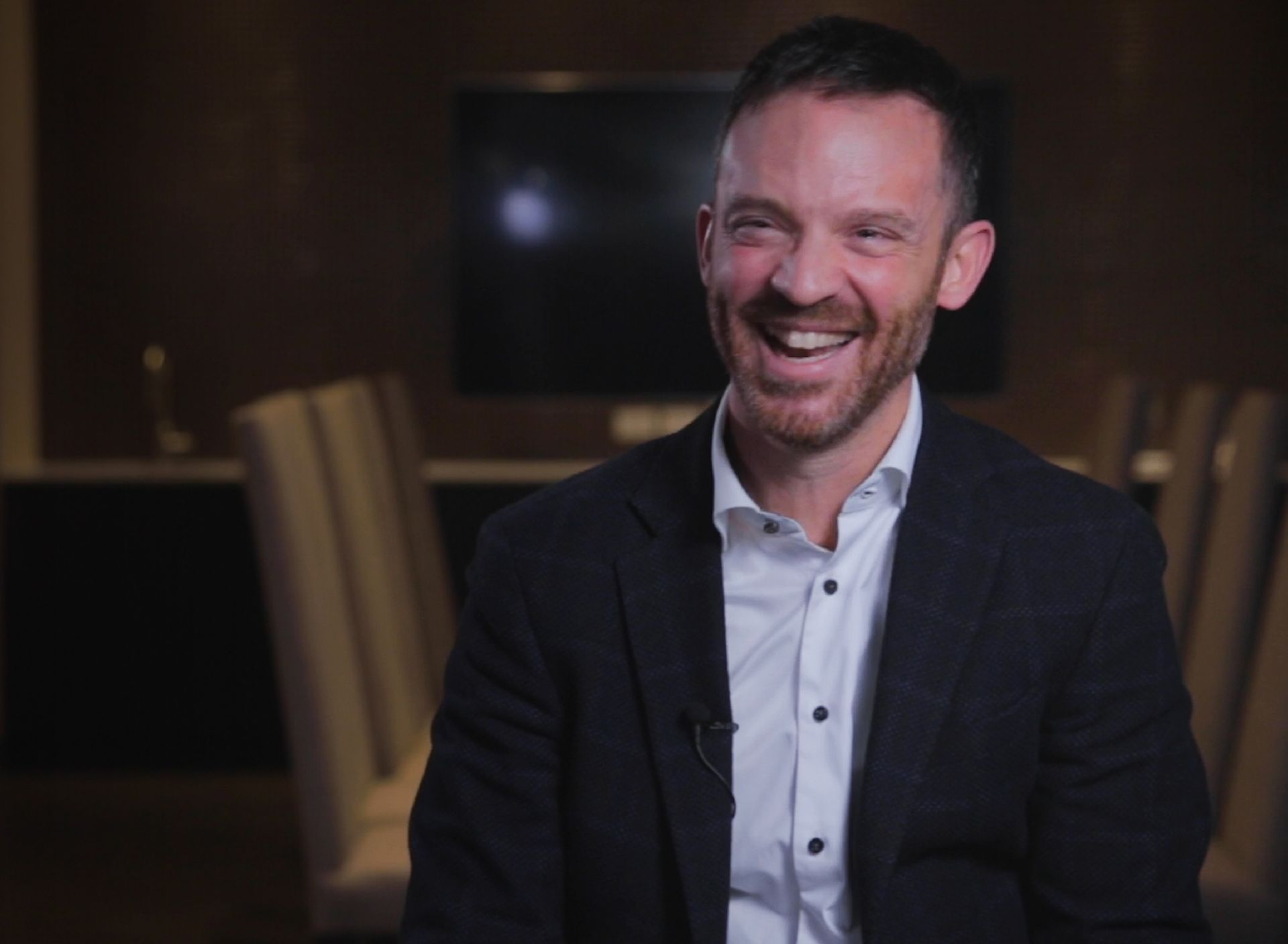 A man in a suit and white shirt is smiling while sitting in a chair.