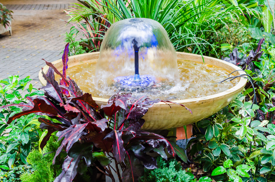 A fountain is sitting in the middle of a garden surrounded by plants.