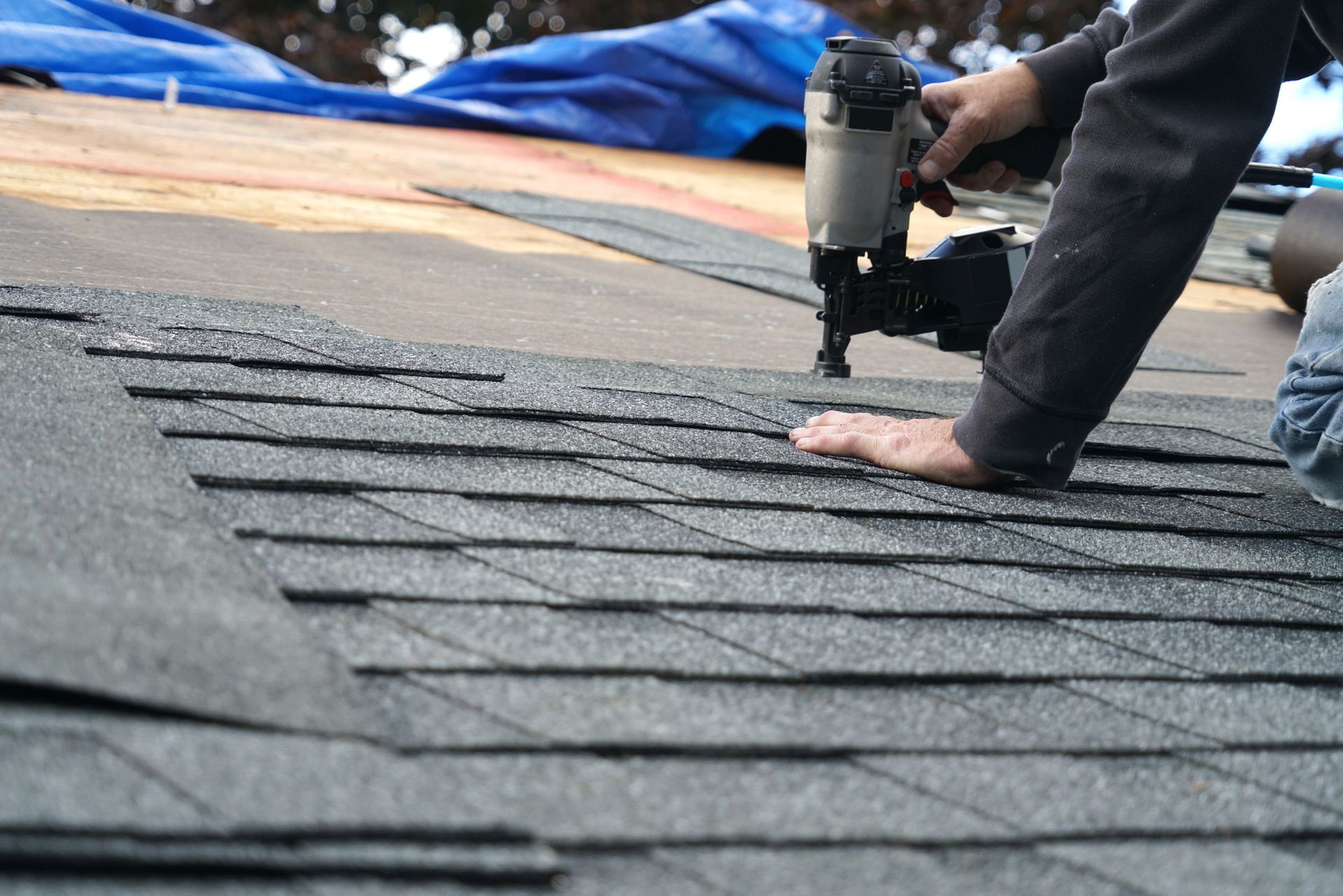 A person using a pneumatic nail gun to install grey asphalt shingles on a residential roof.