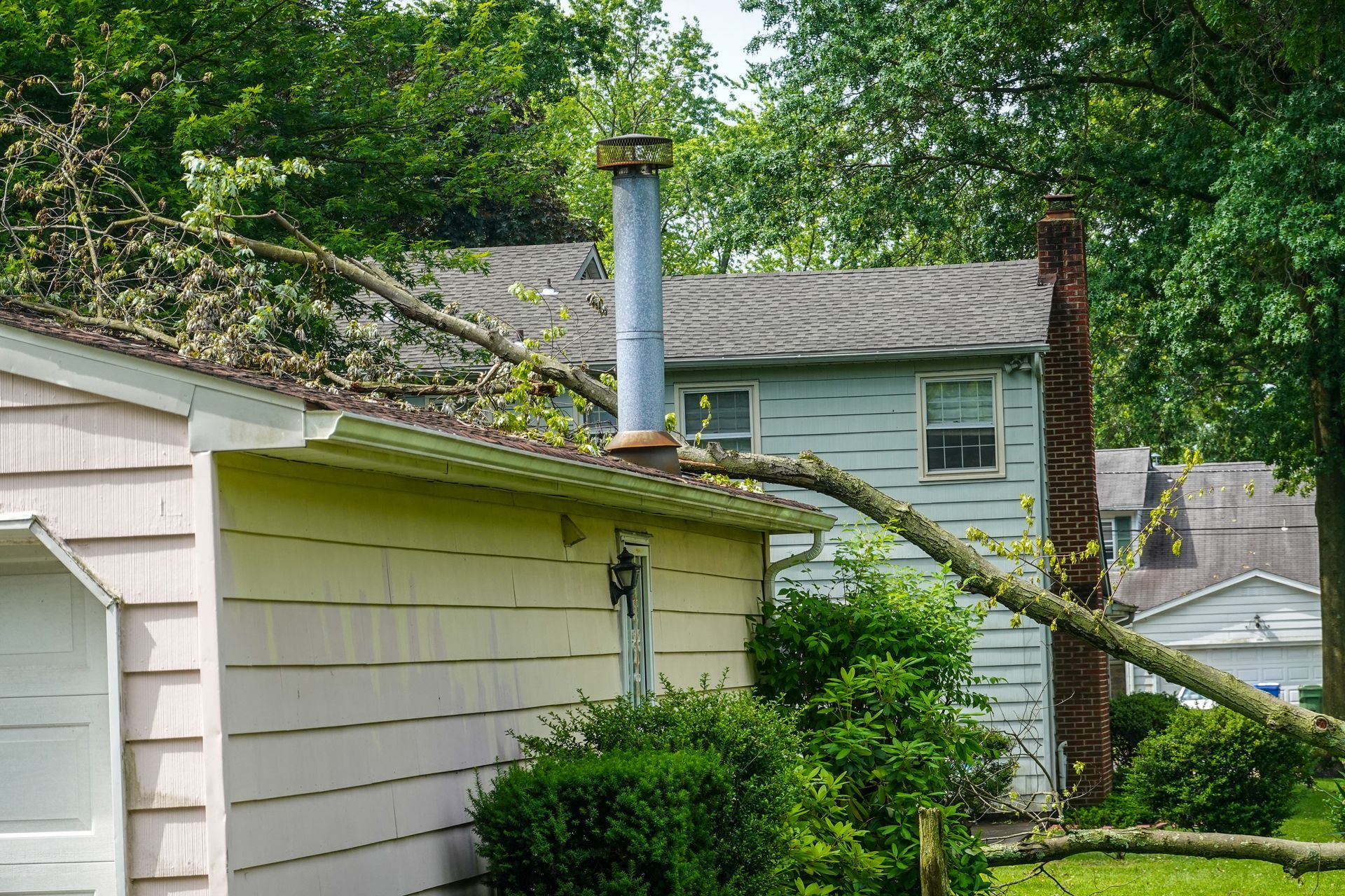 A tree limb has fallen onto the roof of a light pink garage, resting against a metal chimney pipe.