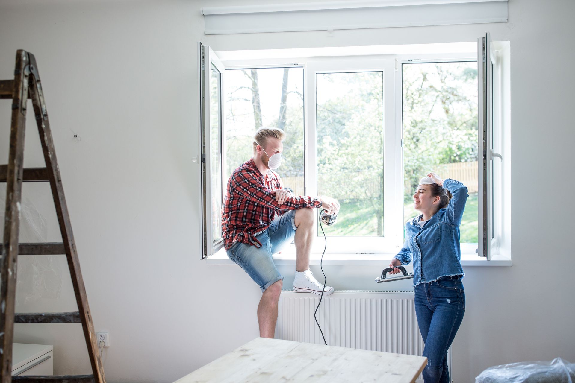 Two people replacing a window during a home renovation, ladder and tools visible.