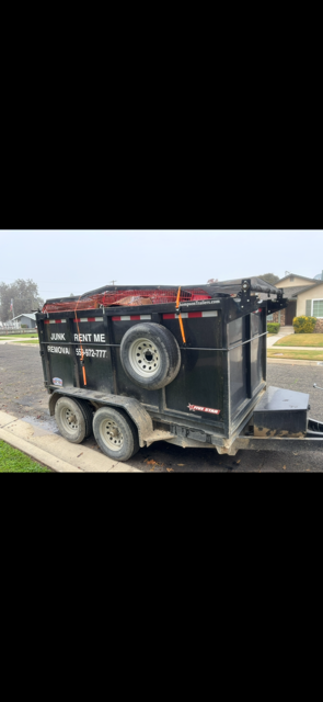A black trailer on the side of a road, with two wheels and spare tire attached. The trailer appears to be full.