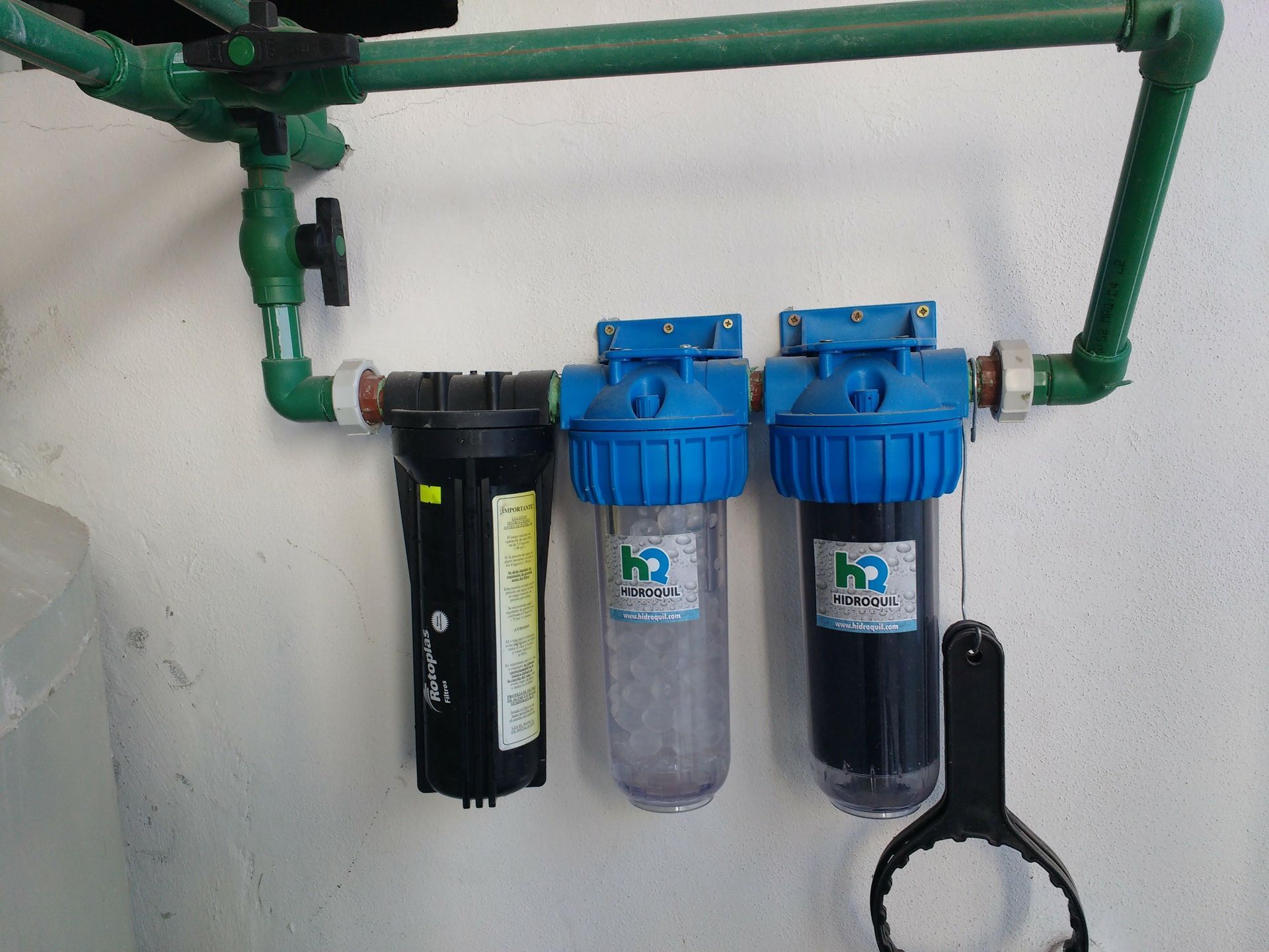 Tres filtros de agua instalados en la pared, conectados por tuberías verdes. Una llave inglesa negra cuelga cerca.