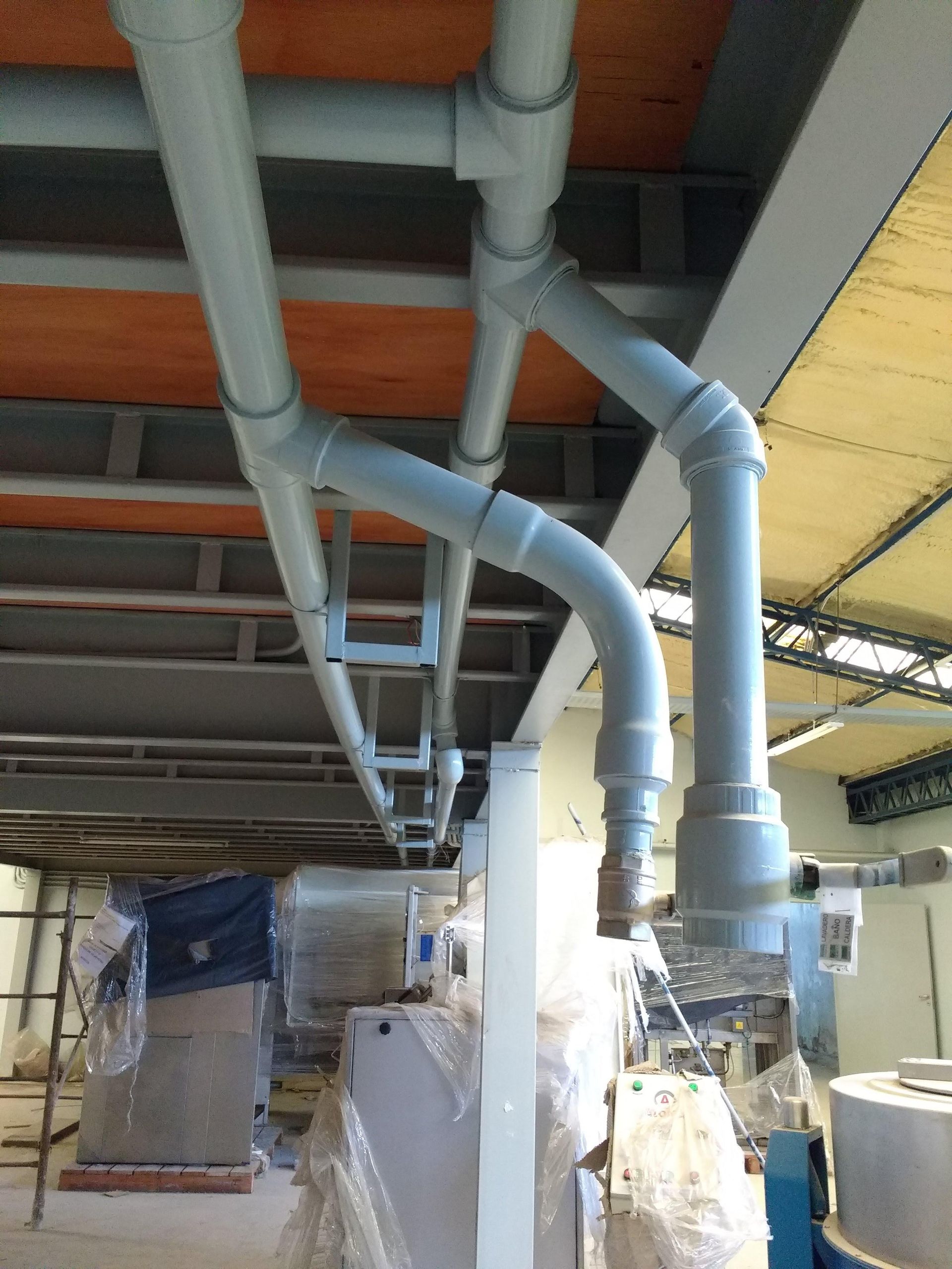 Tuberías grises contra un techo con vigas metálicas y maquinaria debajo.