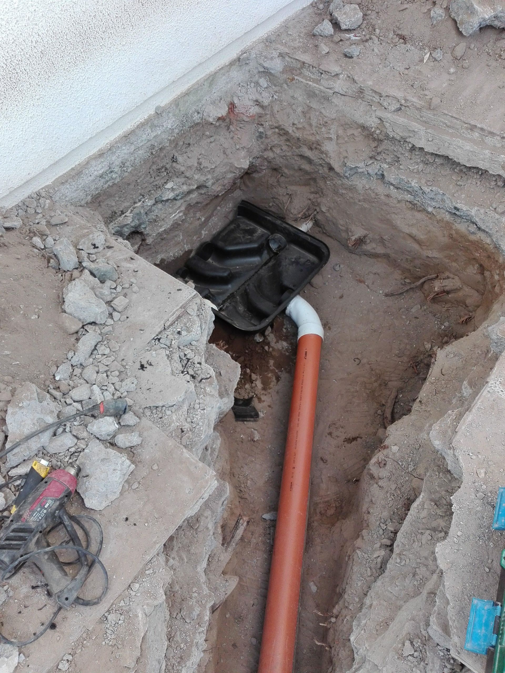 Zanja en la tierra con tubería naranja, codo blanco y recipiente de plástico negro, cerca de un edificio.