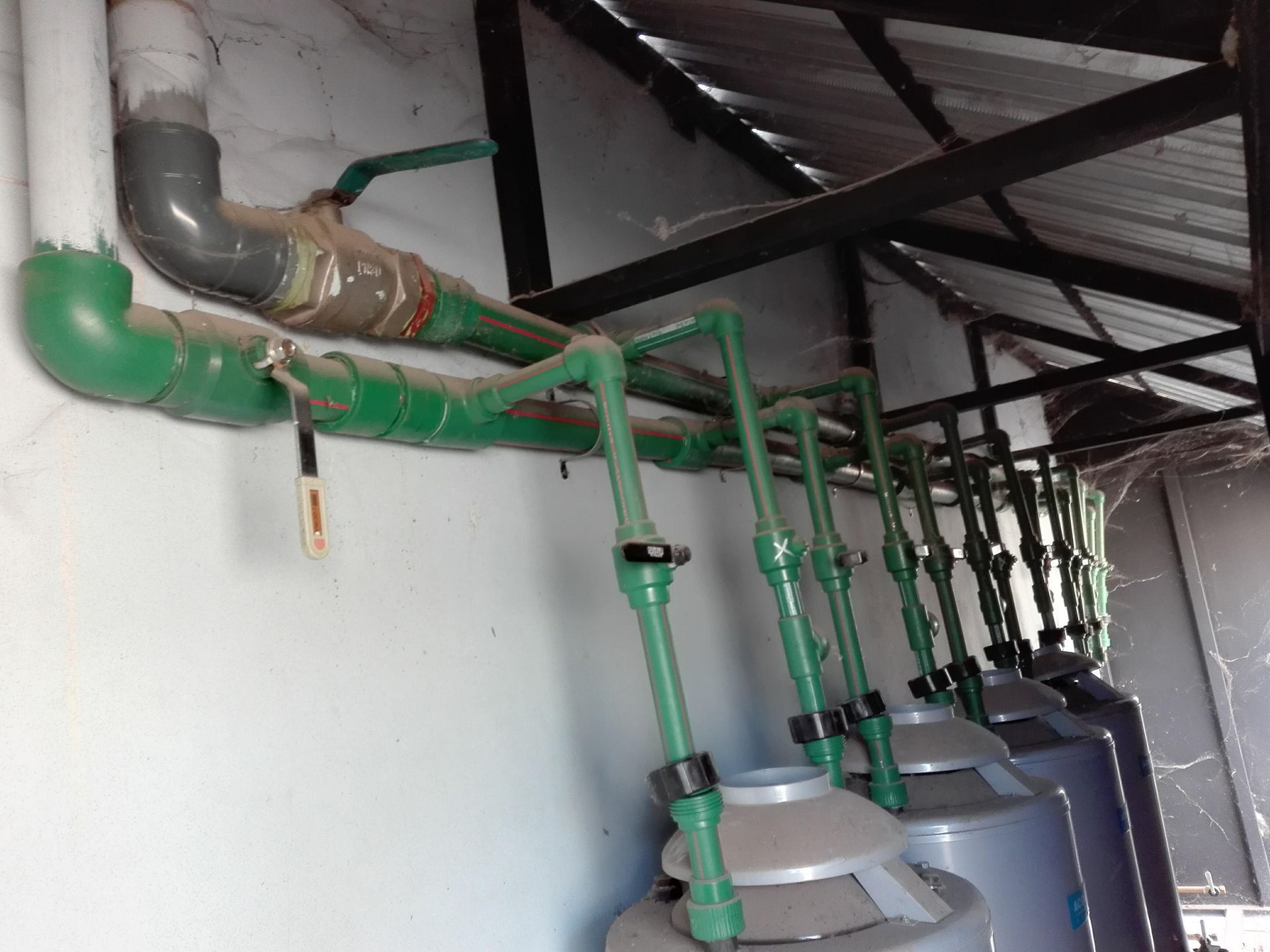 Tuberías, válvulas y depósitos cilíndricos fijados a una pared bajo un techo metálico, posiblemente para uso industrial.