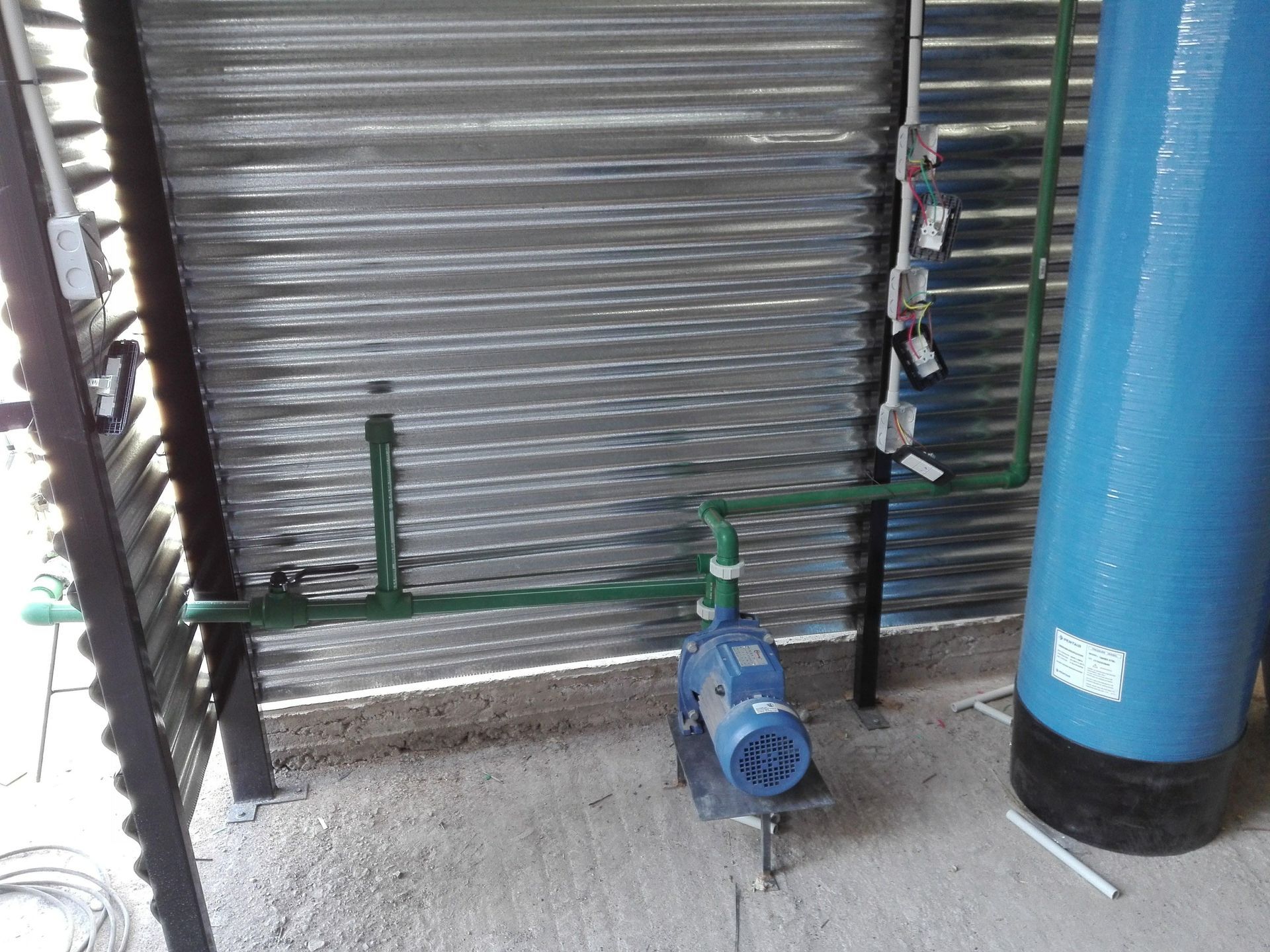 Bomba de agua, tuberías y tanque azul cerca de una pared de metal corrugado.