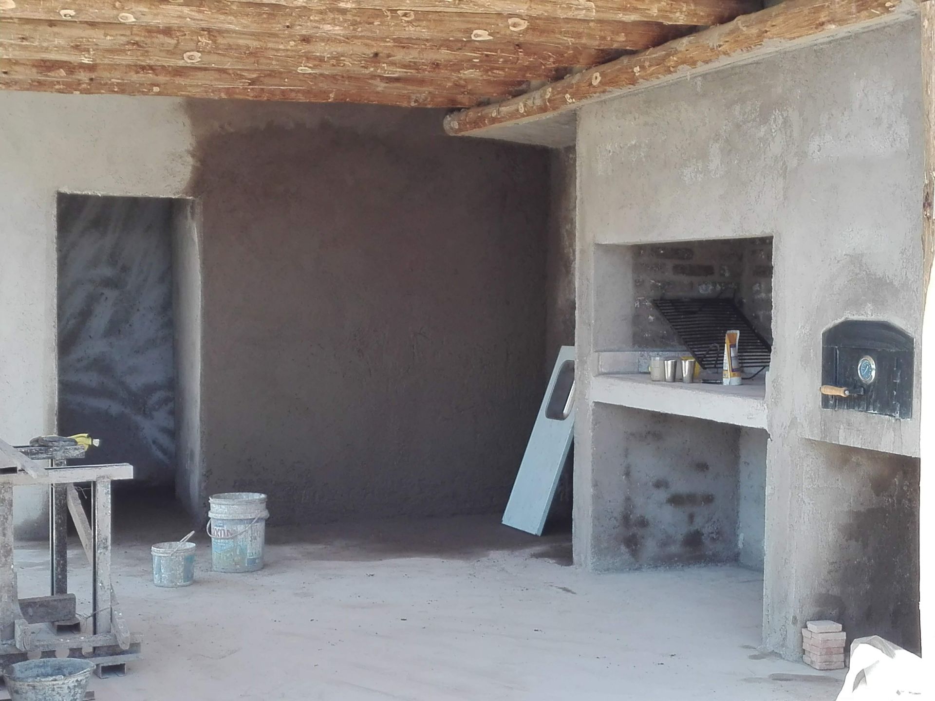 Espacio interior en construcción, con chimenea, horno, puerta, paredes de hormigón y techo de madera.
