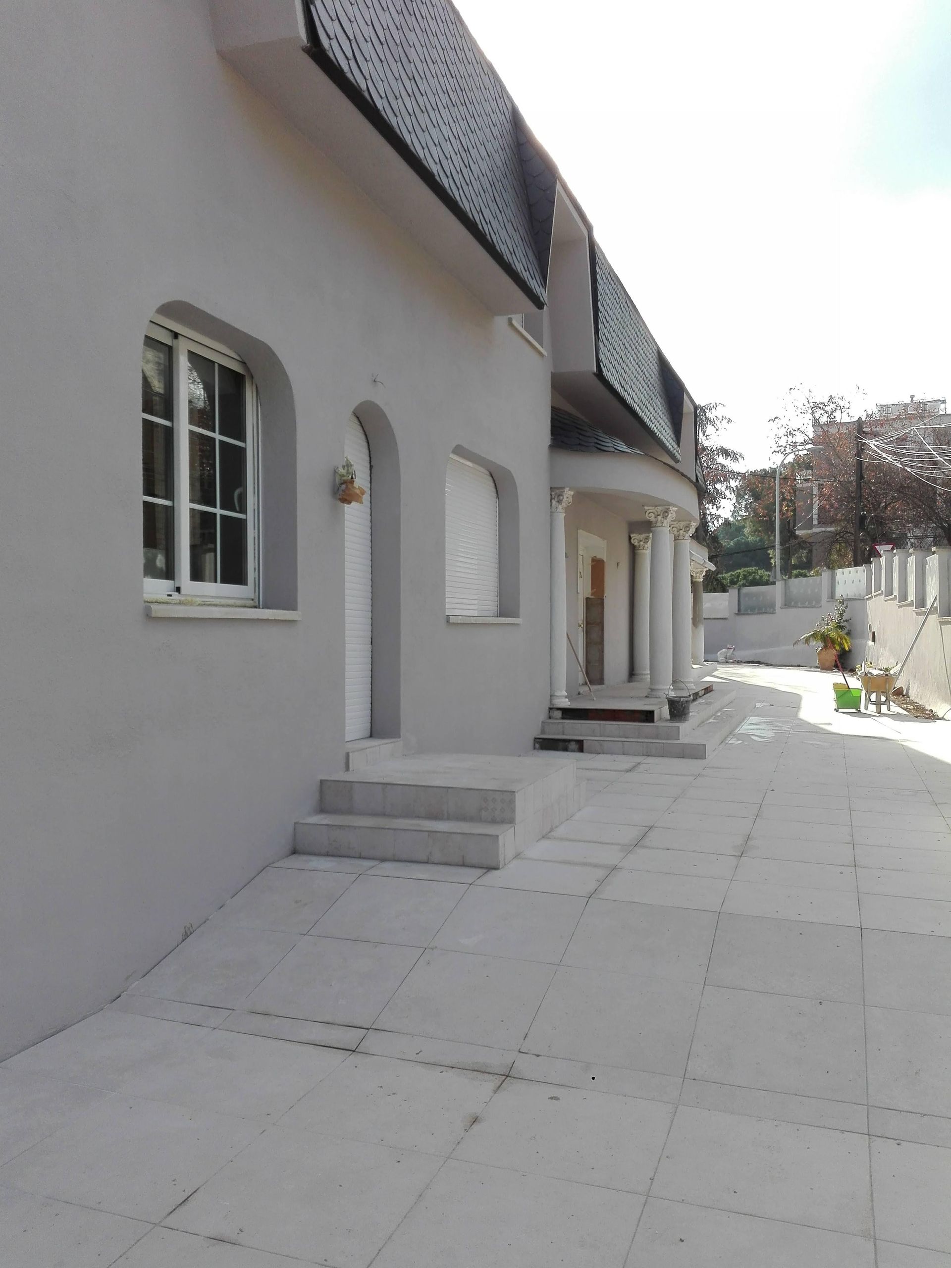 Edificio gris con puerta y ventanas arqueadas, escalones y columnas sobre un patio pavimentado.