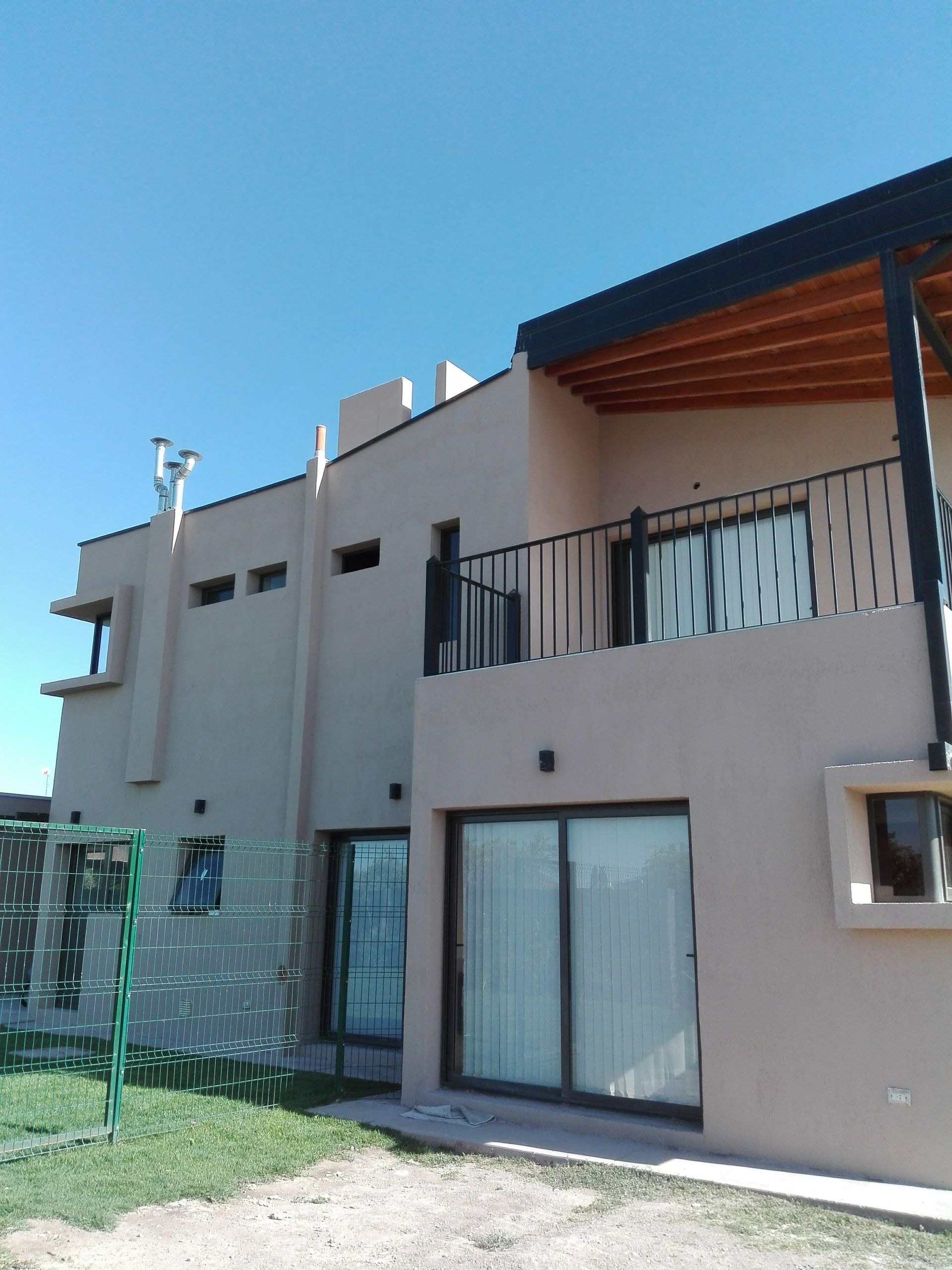 Casa beige de dos plantas con barandilla negra y puertas correderas de cristal bajo un cielo azul brillante.