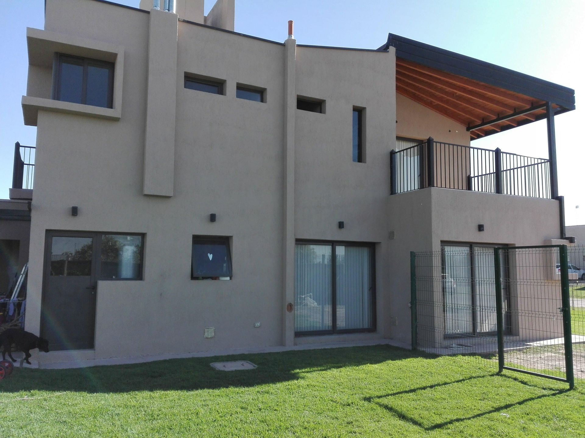 Casa de dos plantas color beige con césped verde, balcón y barandilla negra bajo un cielo soleado y despejado.