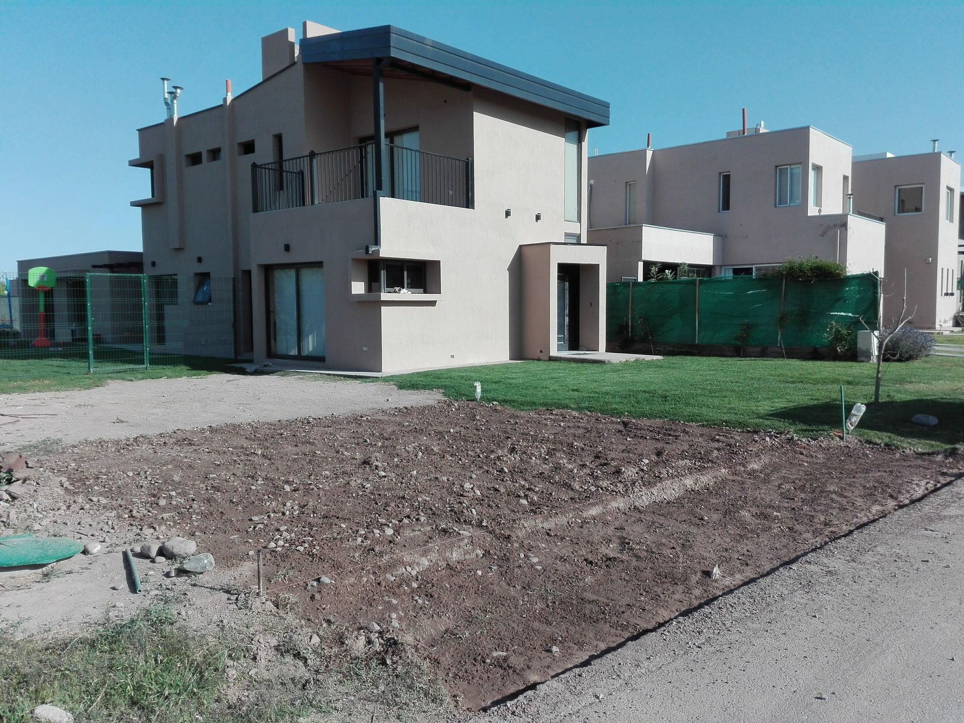 Casa de dos plantas color beige con césped y un trozo de tierra delante; cielo azul despejado.