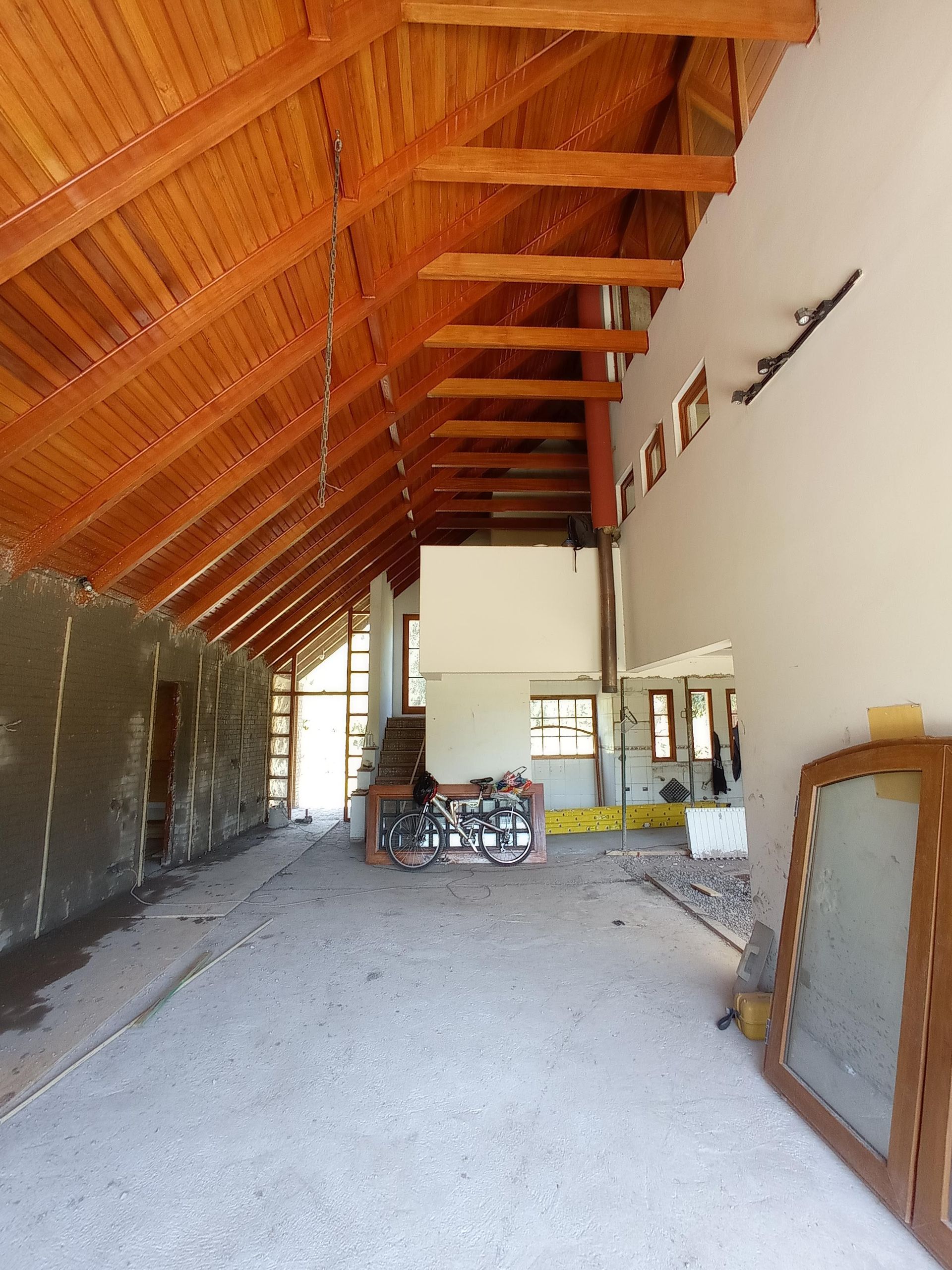 Obra en construcción interior con vigas y techo de madera, suelo de hormigón, paredes blancas.