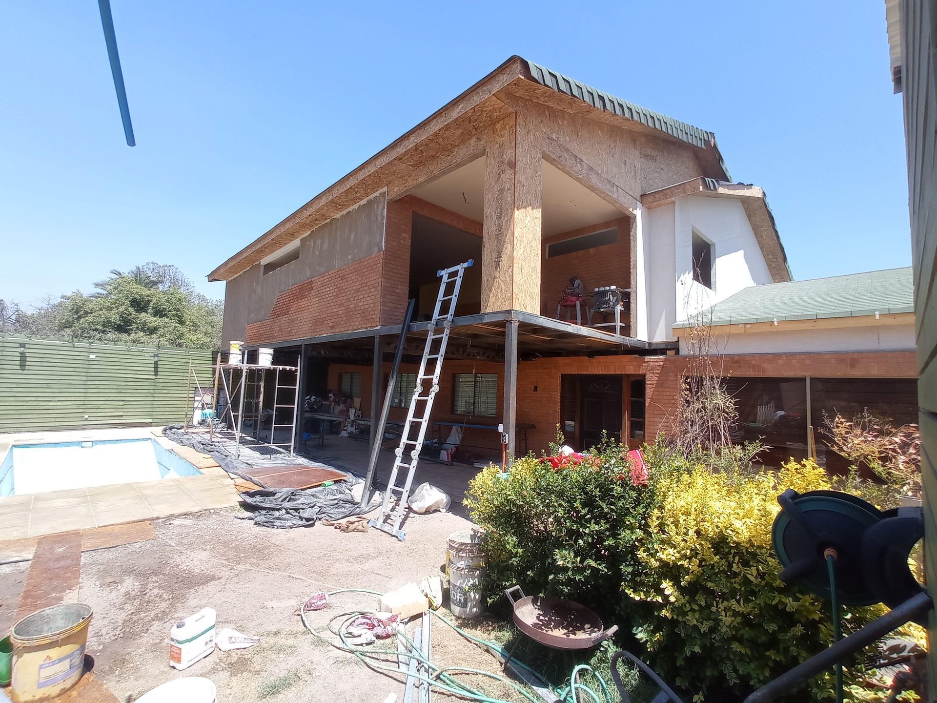 Casa en construcción con piscina, paredes de madera, escalera y vegetación.