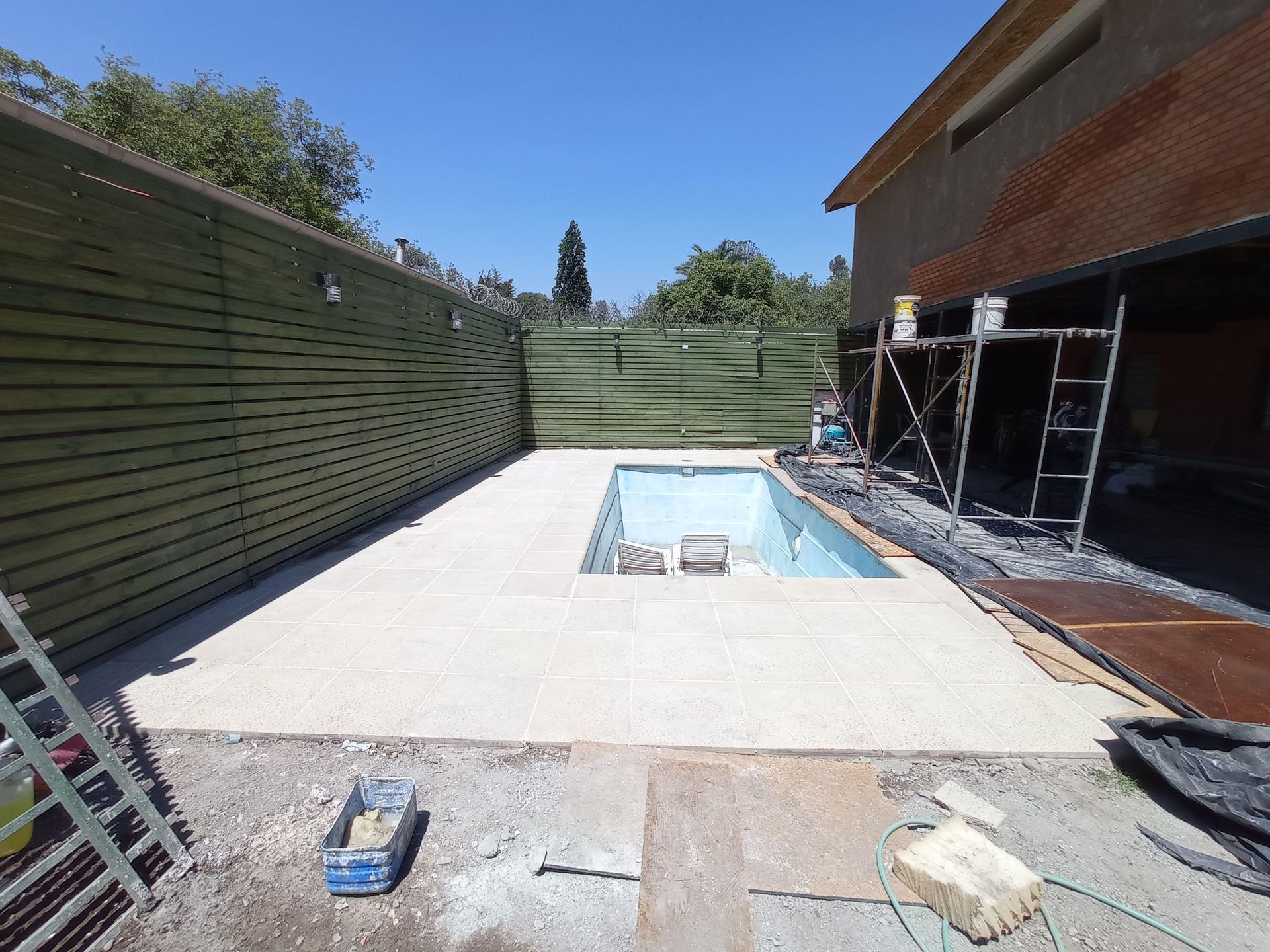 Piscina en construcción junto a una valla de madera y una casa; borde de hormigón.
