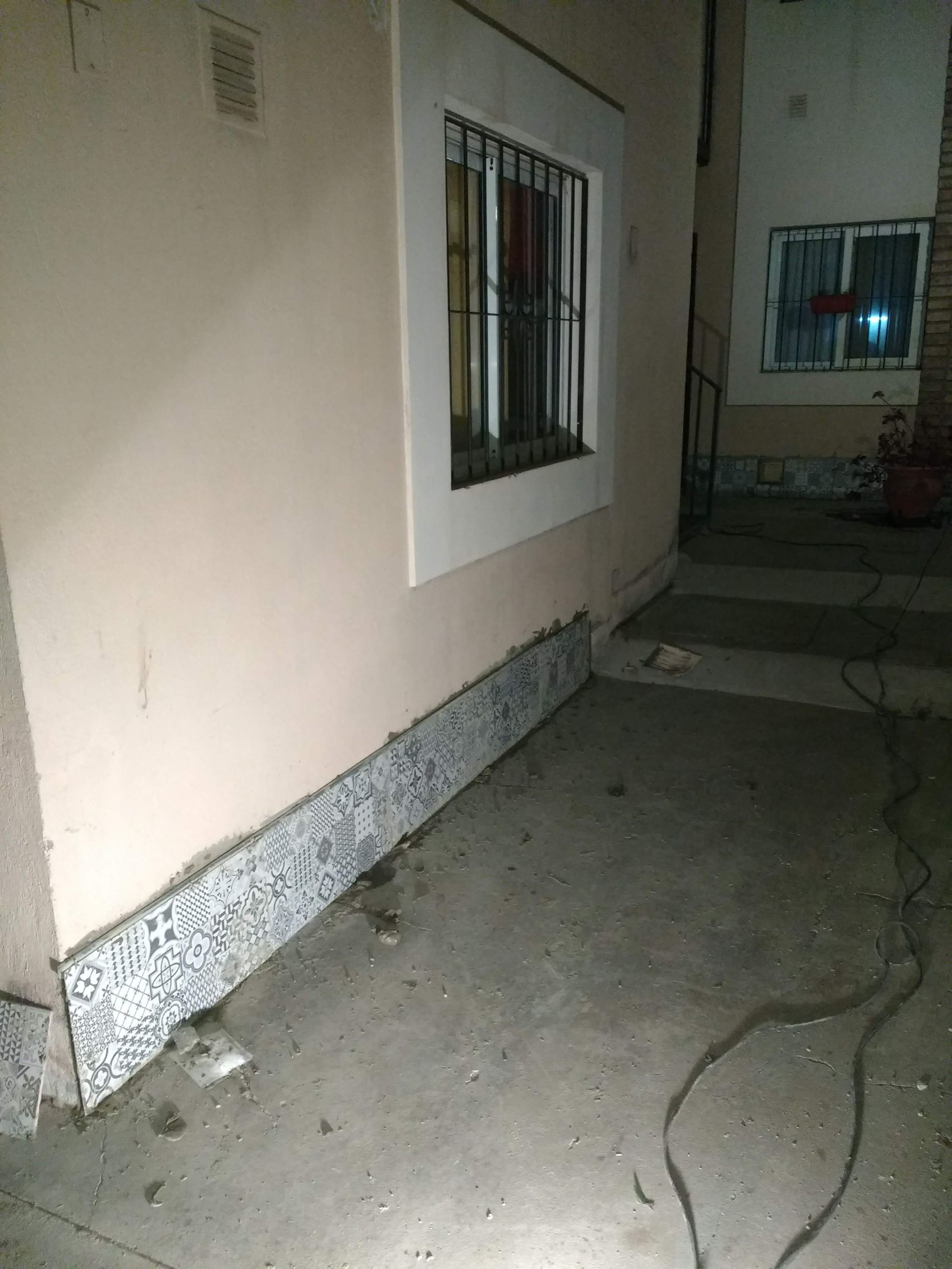 Esquina exterior del edificio; pared beige, ventana enrejada, base texturizada, suelo de hormigón, oscuro.