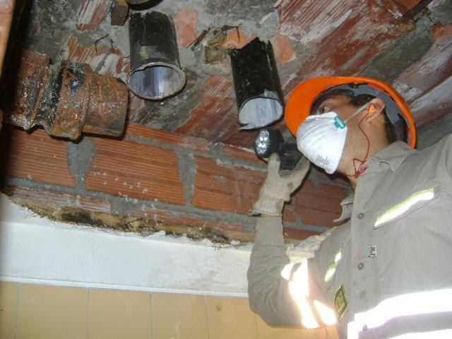 Trabajador con equipo de seguridad inspeccionando tuberías con una linterna, cerca de ladrillos y techo.