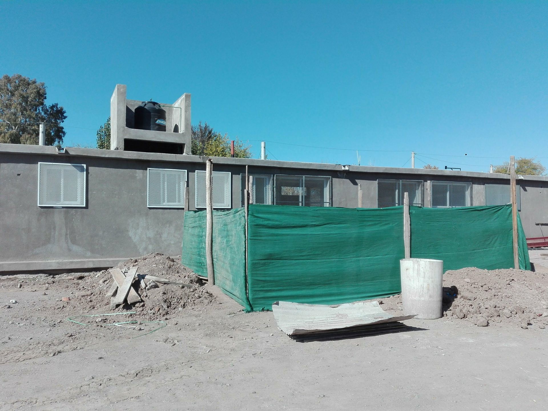 Exterior de un edificio gris con ventanas, una valla de malla verde y materiales de construcción bajo un cielo azul.