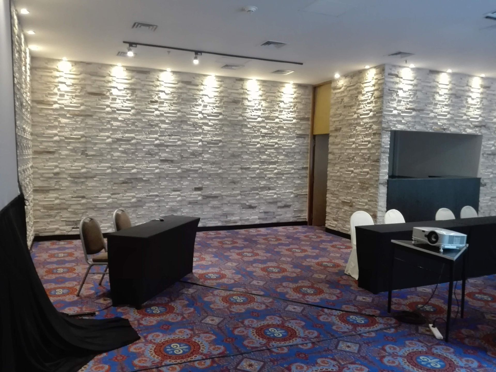 Sala de conferencias con paredes de piedra texturizada, mesas, sillas, proyector y pantalla.