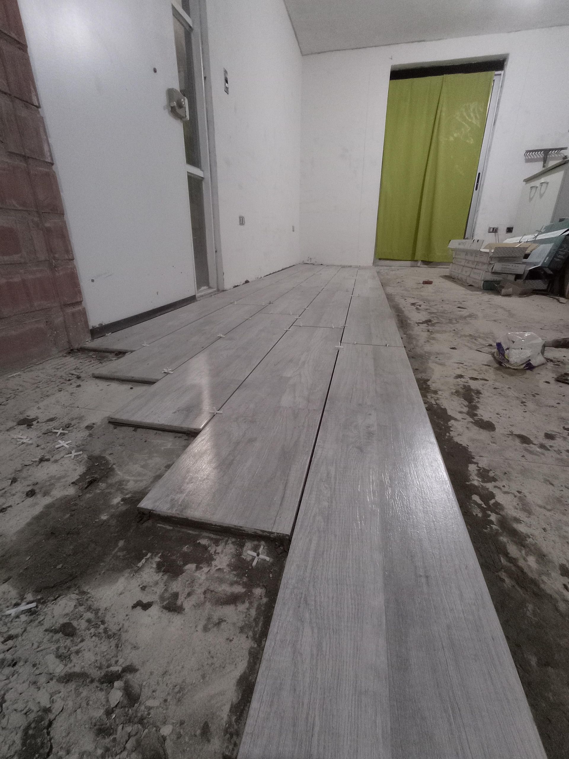 Instalación de suelo de tablones grises en curso; habitación con paredes blancas, puerta y cortina verde.