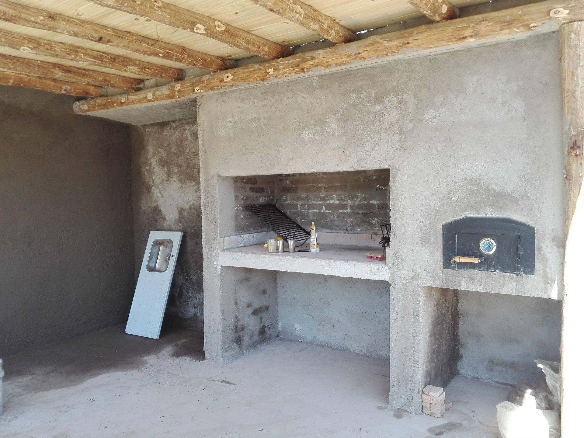 Cocina exterior con estructura de cemento que incluye parrilla, horno y mostrador; bajo un toldo de madera.