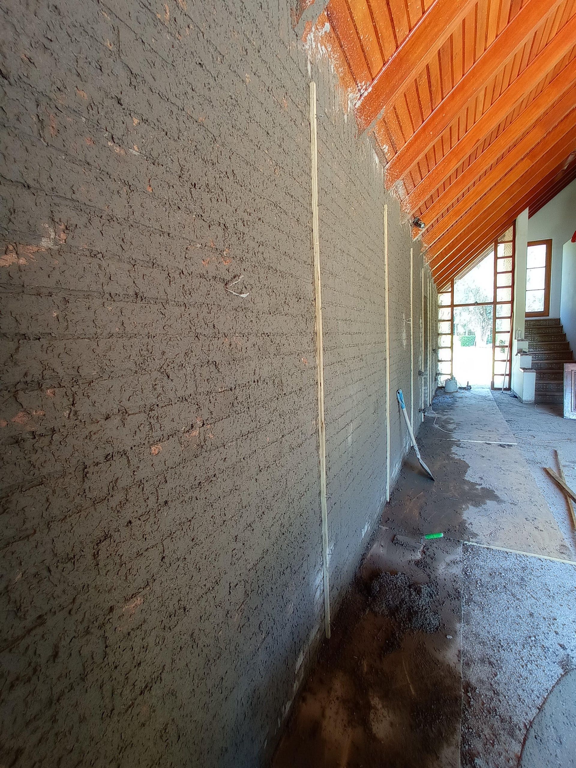 Pared de estuco gris en construcción, con textura visible. Adyacente a un interior sin terminar y techo de madera.