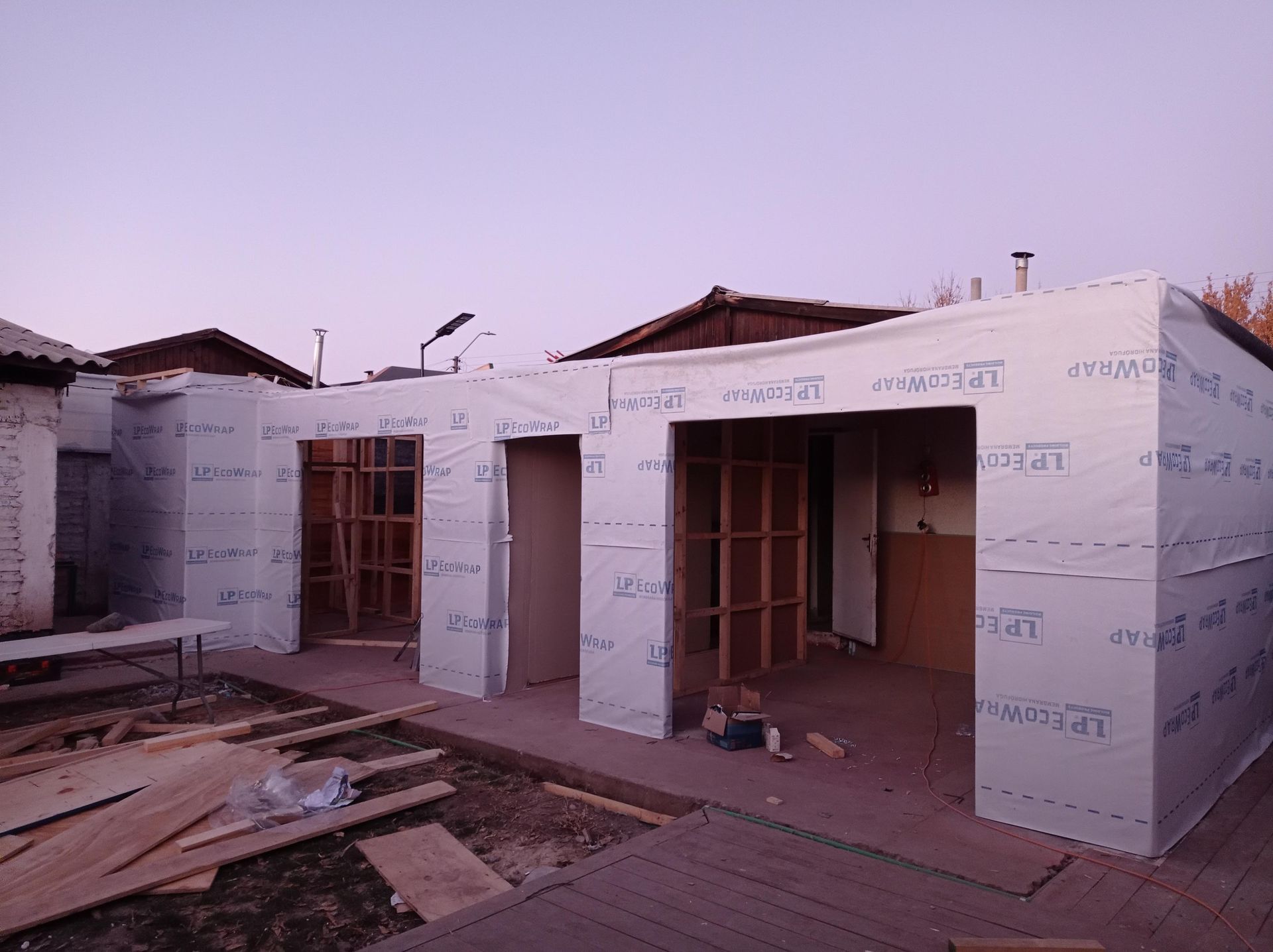 Construcción del exterior de un edificio con estructura de madera y revestimiento blanco impermeable. Ambiente al atardecer.