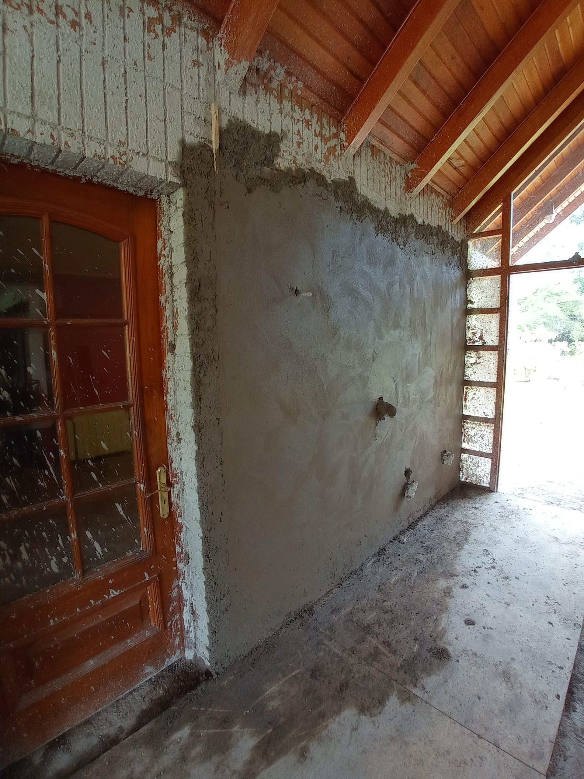 Muro exterior con estuco parcialmente aplicado, junto a una puerta de madera y un porche.