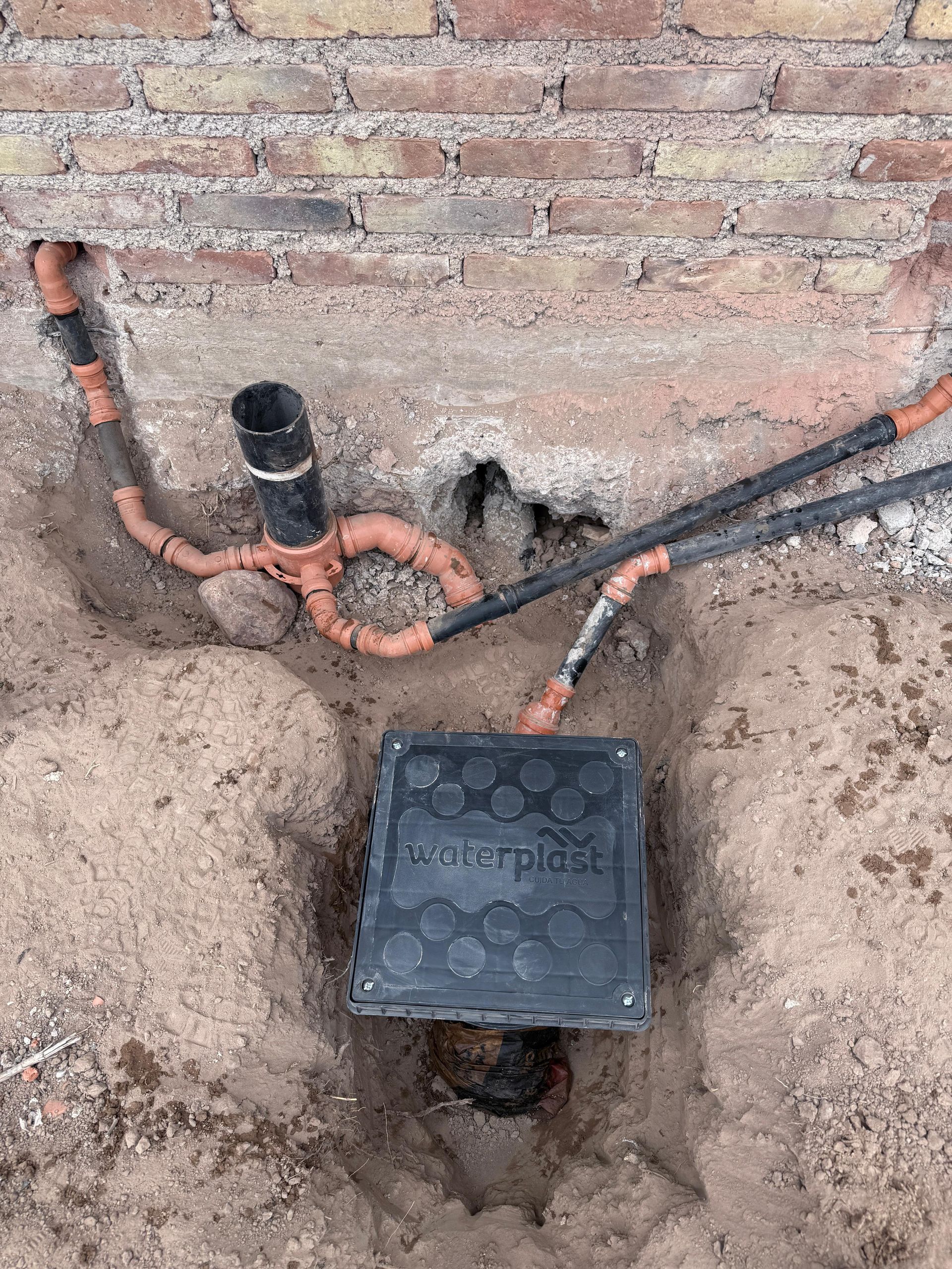 Caja de servicios negra con tuberías naranjas y negras conectadas en una excavación cerca de una pared de ladrillo.