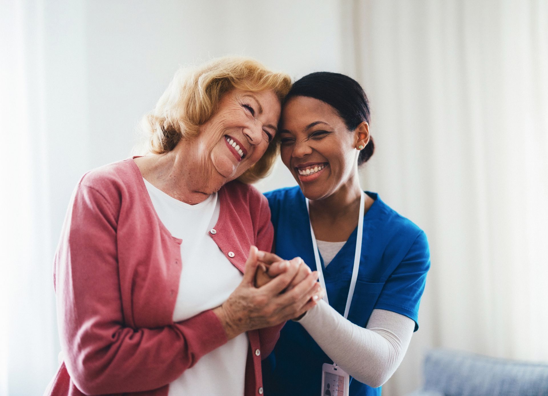 Smiling elderly woman with caregiver; both hold hands, indoor.