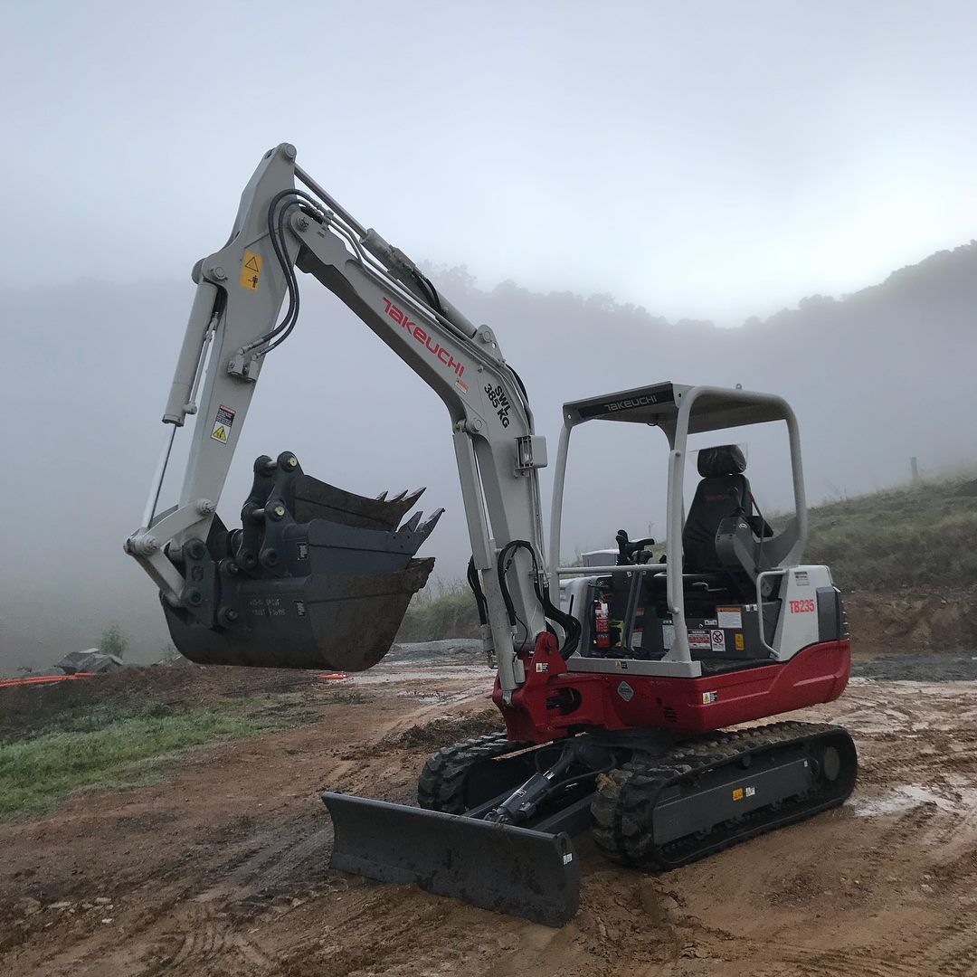 Small Excavator on Muddy Ground With Bucket Raised, Foggy Hillside in Background — Azza's Mini Diggers in Port Macquarie, NSW