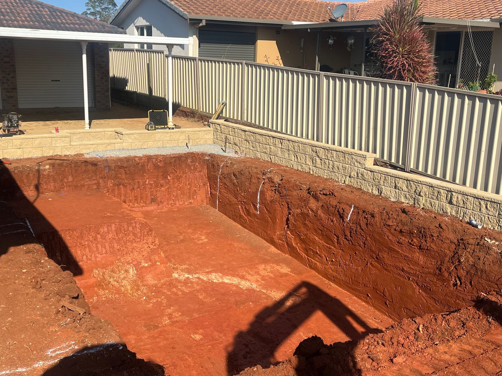 Excavated rectangular hole in red soil, surrounded by a fence and houses. Shadow of excavator visible. — Azza's Mini Diggers in Port Macquarie, NSW