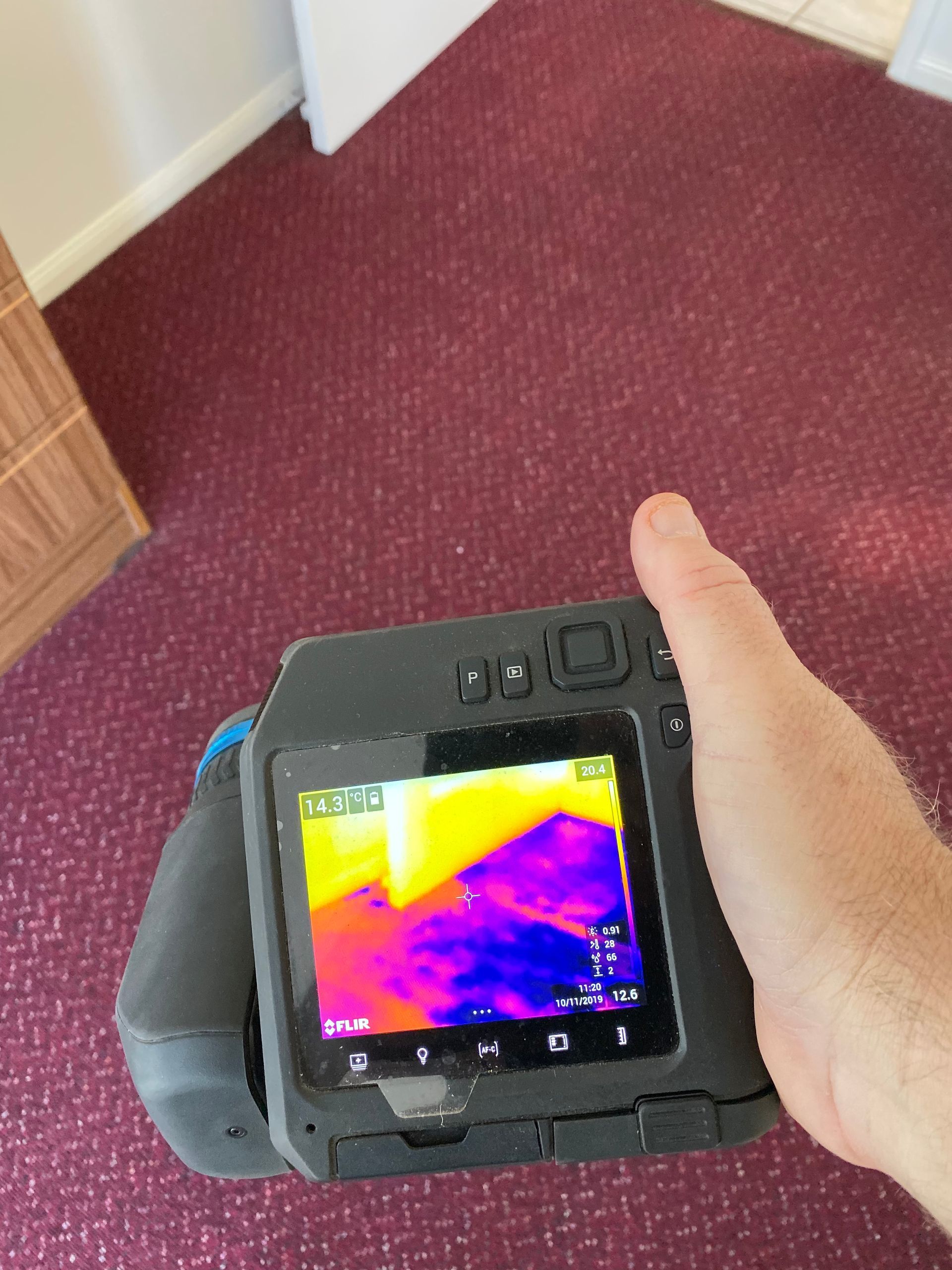Hand holding a thermal imaging camera pointed at a purple carpet. The camera screen shows color gradients.