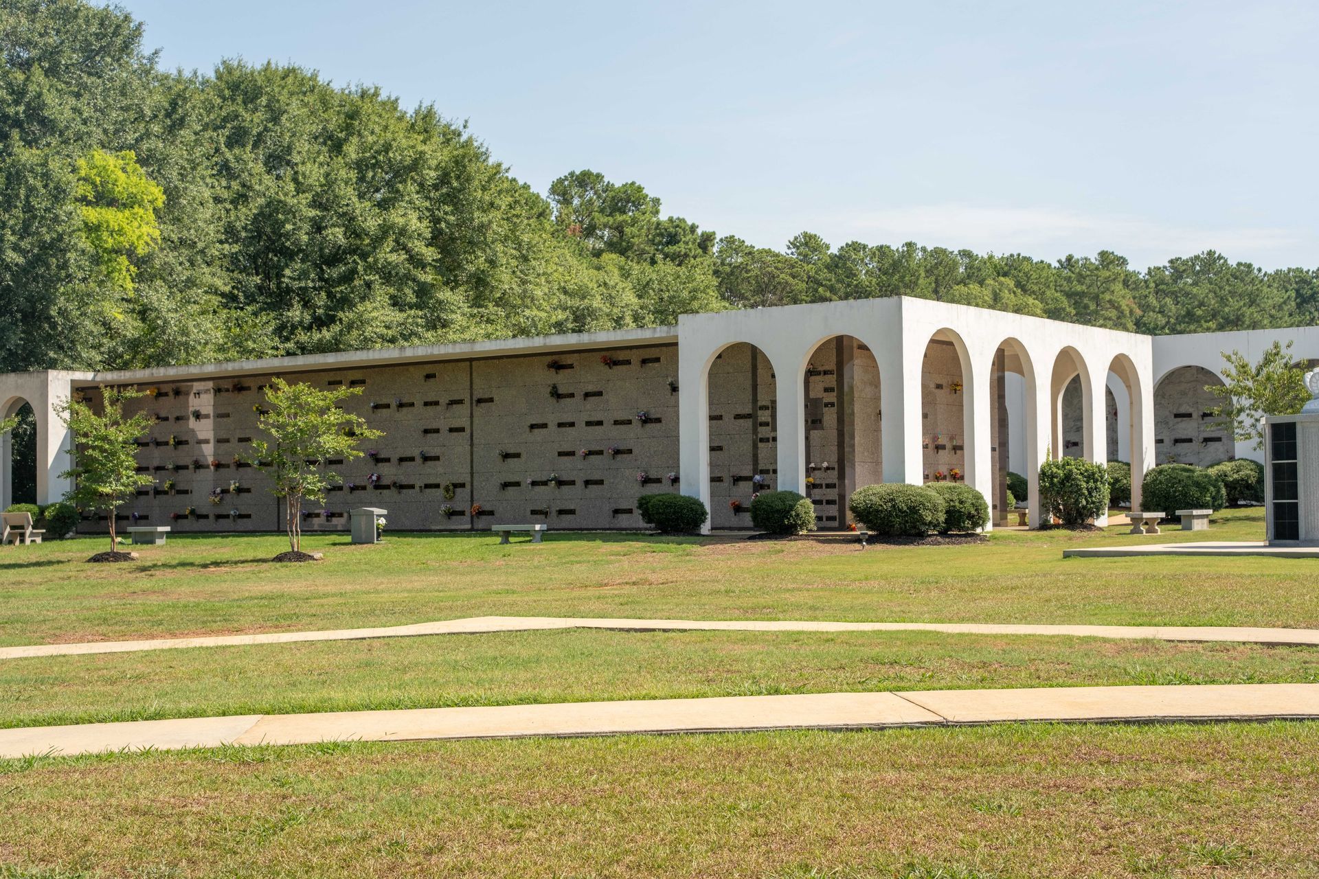 Exterior of a mausoleum, featuring arched entrances and niches with headstones set in the wall, surrounded by grass and trees.