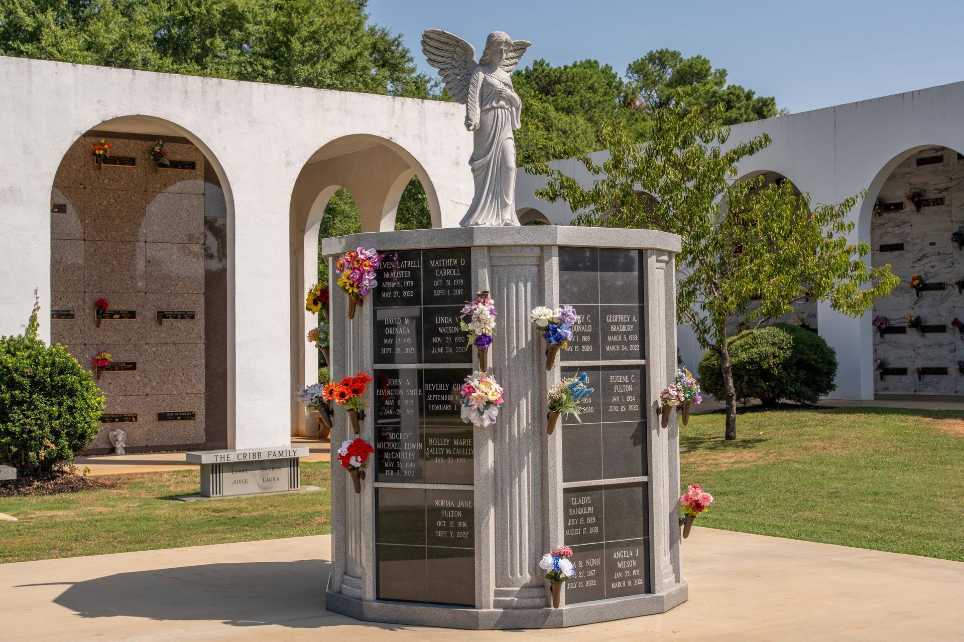 A mausoleum with an angel statue on top, surrounded by columbarium niches, and colorful flower bouquets.