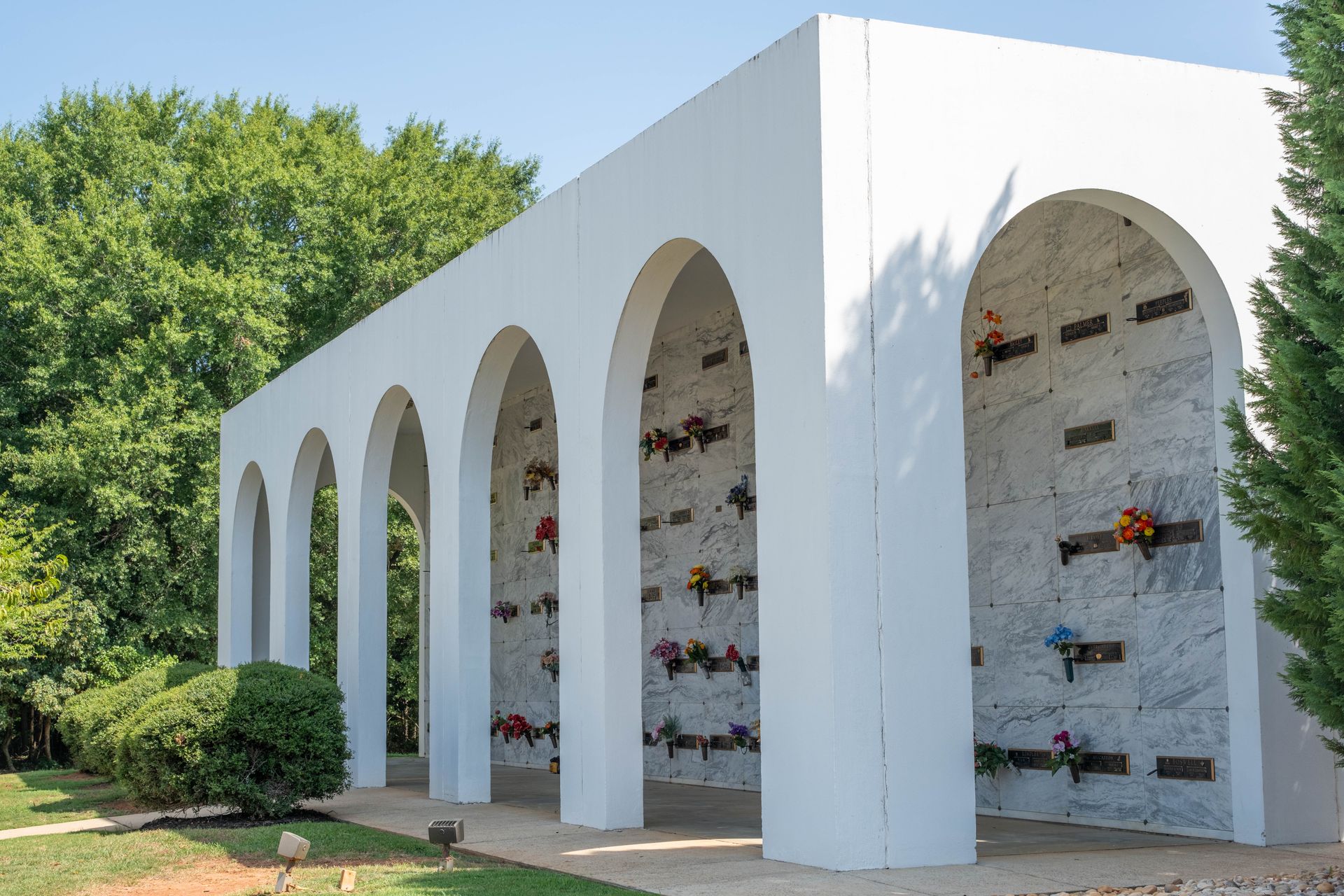 White arched columbarium with niches holding urns; set outdoors, surrounded by trees and greenery.