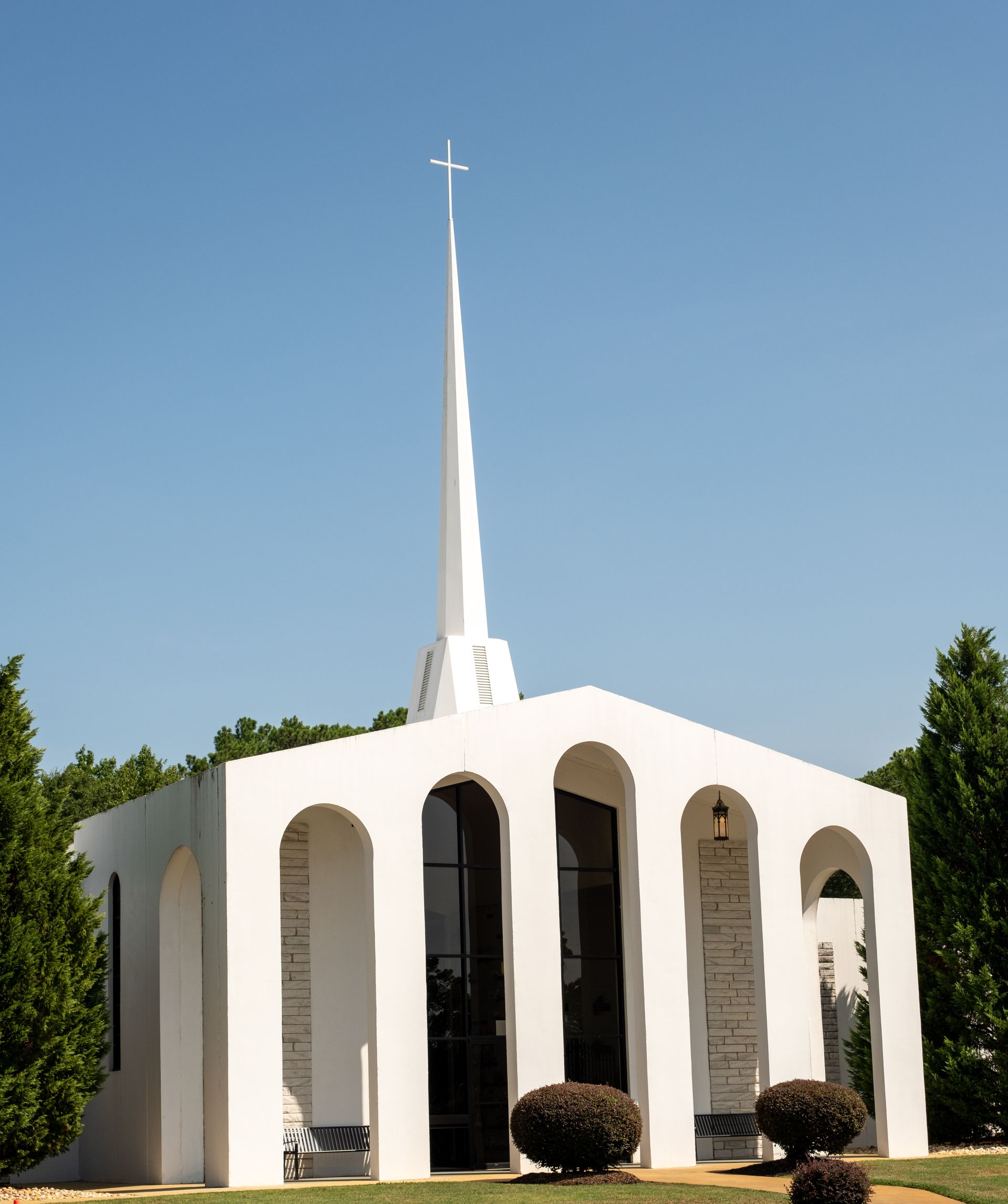 White chapel with tall spire, arched windows, and small bushes under a clear blue sky.