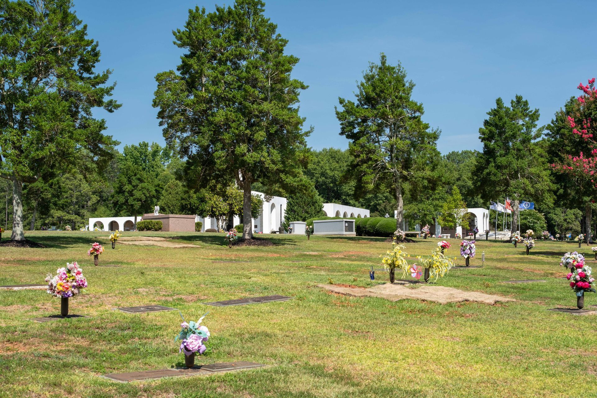 A cemetery with green grass, trees, and white structures on a sunny day. Artificial flowers decorate graves.