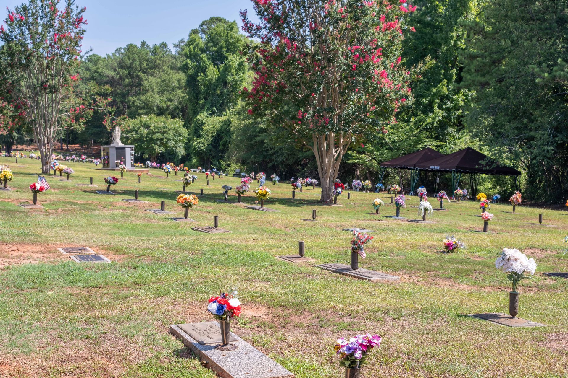 Lawn cemetery with numerous graves marked by headstones and flowers. Trees and a small gazebo visible.