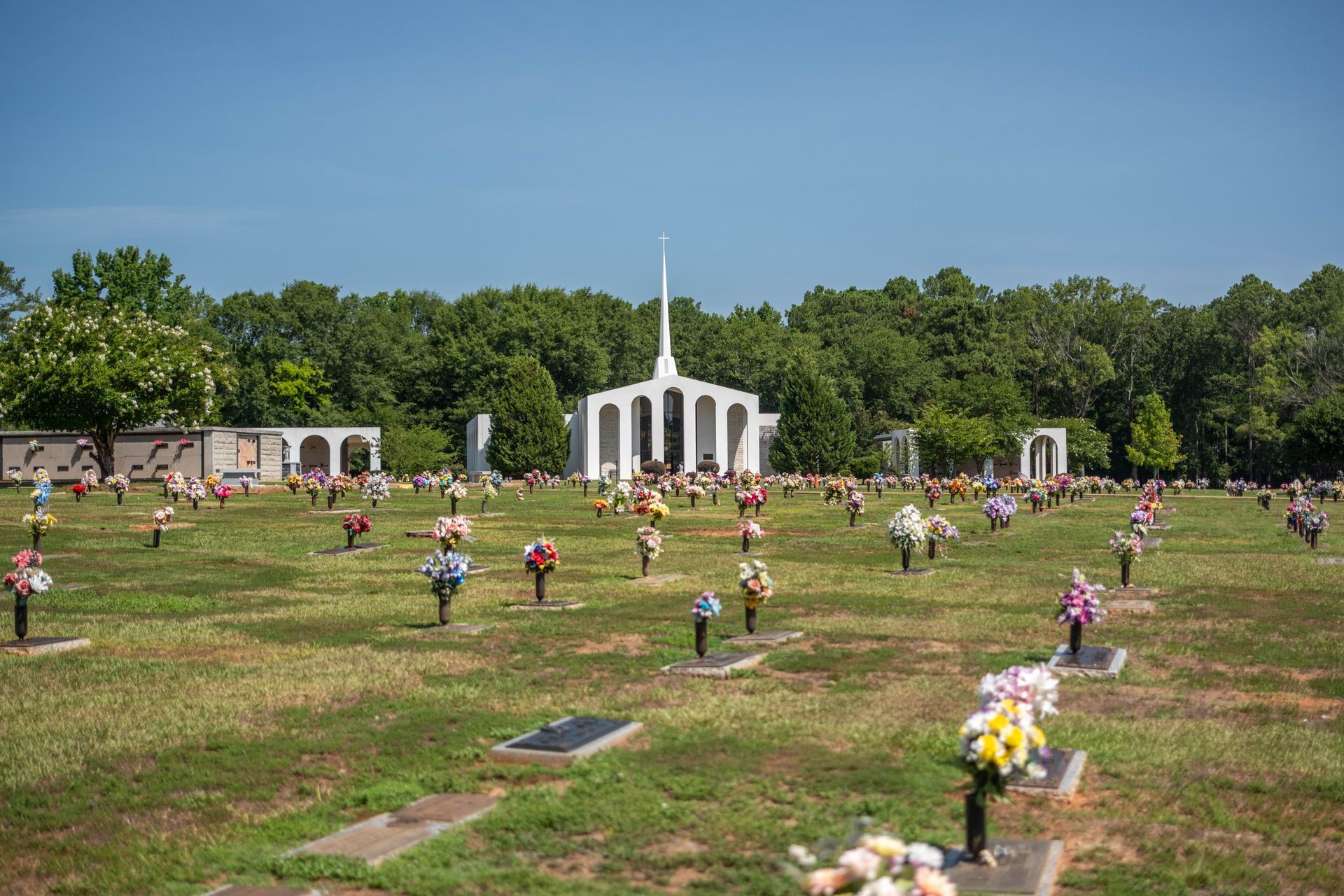 Cemetery with many headstones and colorful flower arrangements in front of a white building with a steeple.
