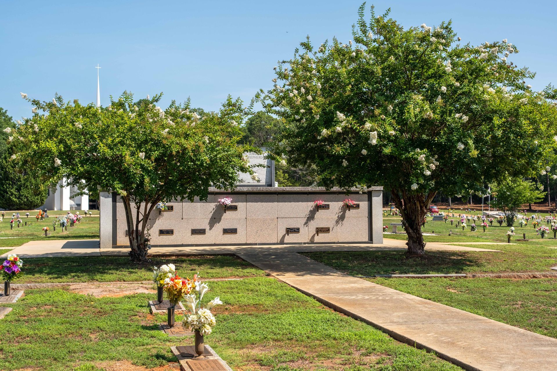 Cemetery scene with a concrete columbarium flanked by green trees and a pathway, with gravestones in the background.