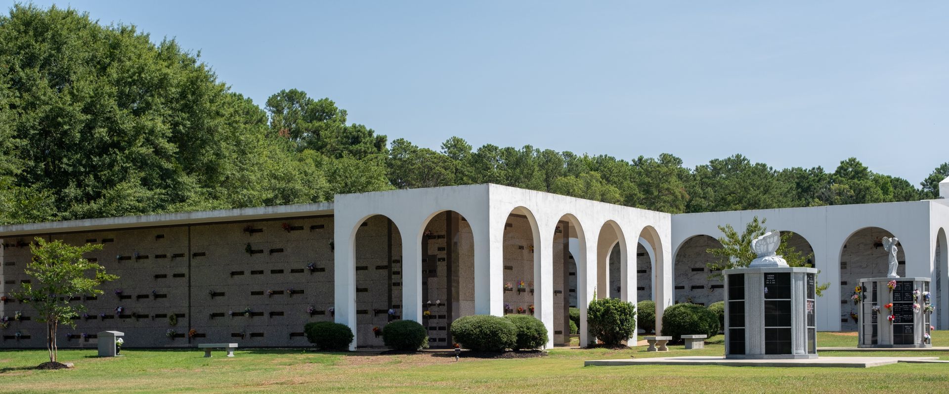 A cemetery with white arched structures and headstones, with a green lawn and trees under a blue sky.
