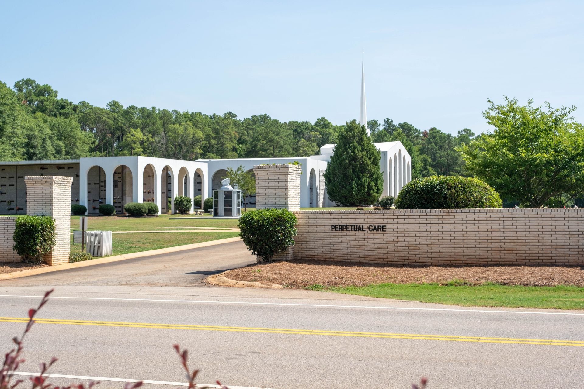 Cemetery entrance with arched structures, grassy areas, and trees under a sunny sky.
